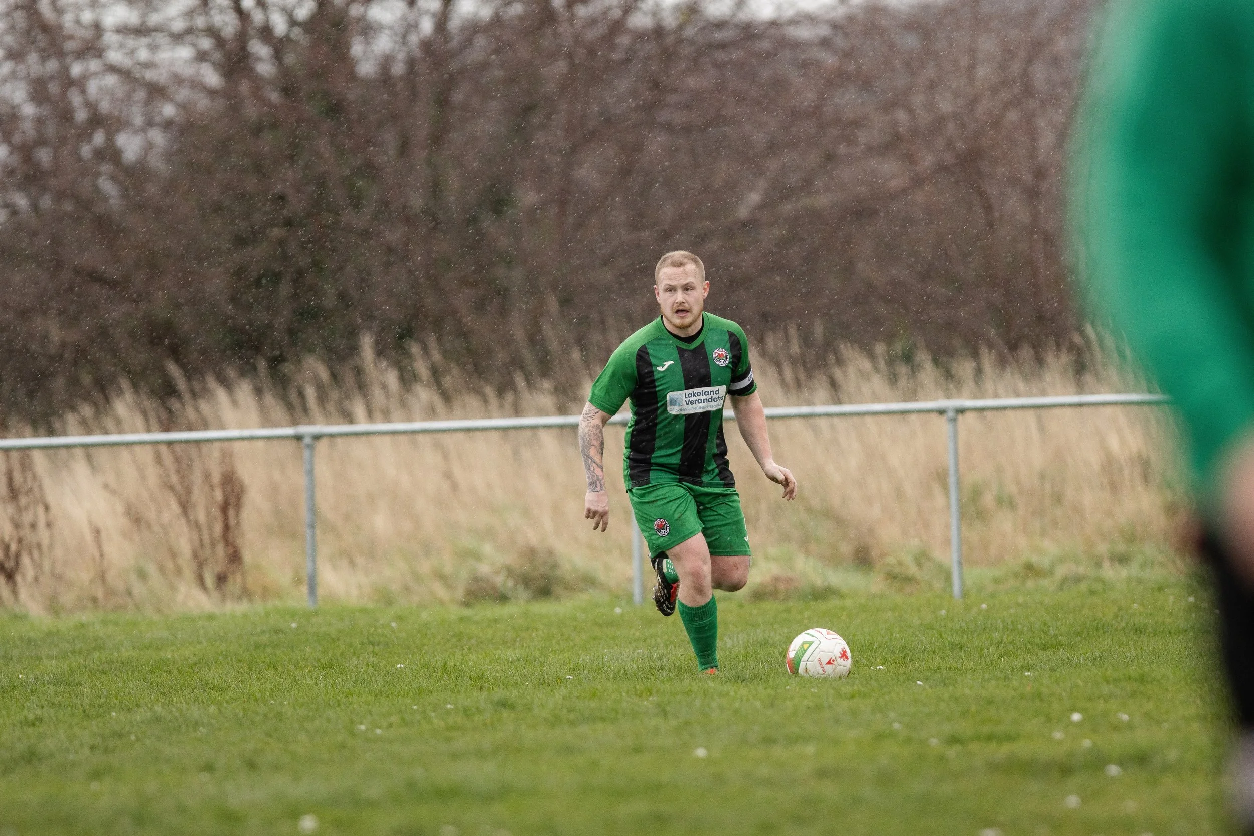 A male soccer player in a green and black uniform is on a grassy field, preparing to kick a white soccer ball. The player has tattoos on his right arm and is wearing a captain's armband. Trees and a metal fence are in the background.