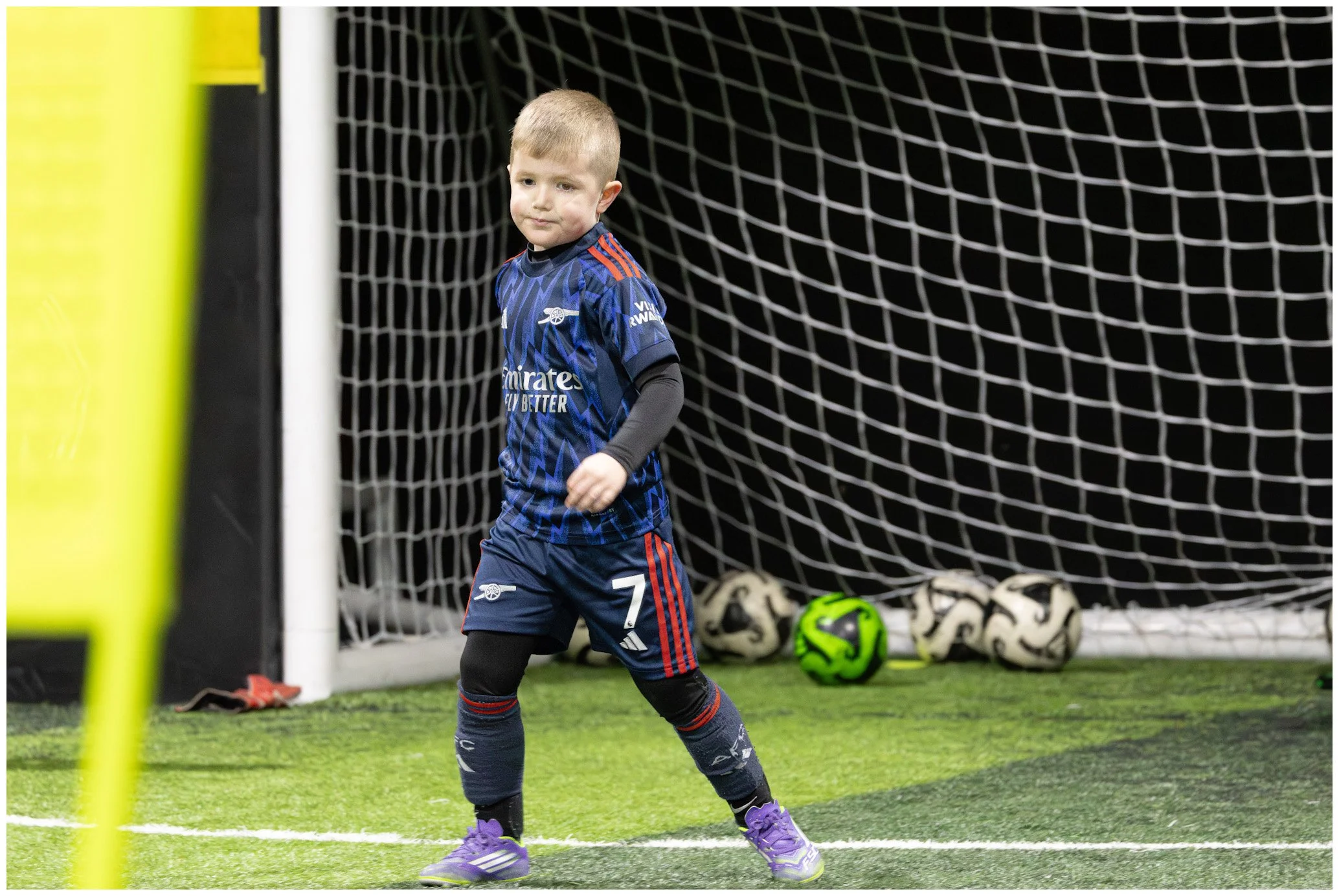 A young boy dressed in a blue soccer jersey with red accents, standing in an indoor soccer field near a goal, with soccer balls in the background.