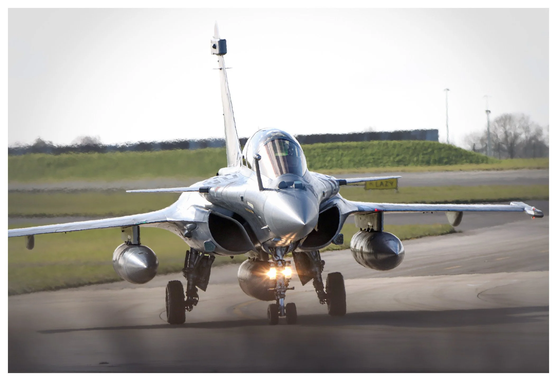 A silver fighter jet taxiing on a runway with landing gear, surrounded by green grass and an overcast sky.