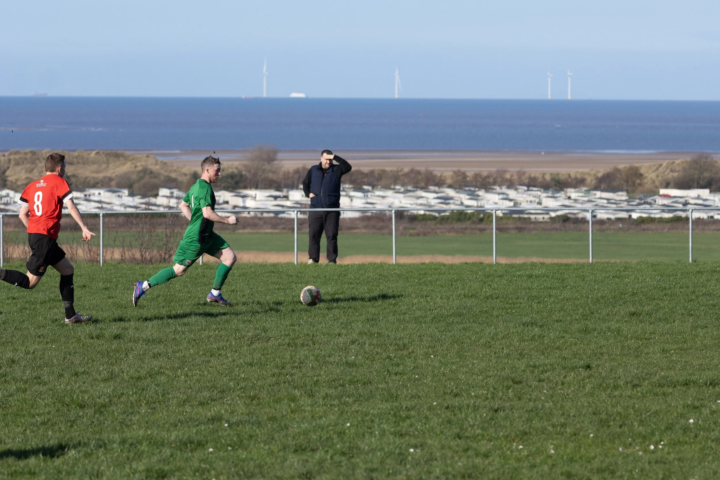 Two young men playing soccer on a grassy field, one wearing a green uniform and the other in red, with a spectator watching behind a fence and a coastal landscape in the background with wind turbines.