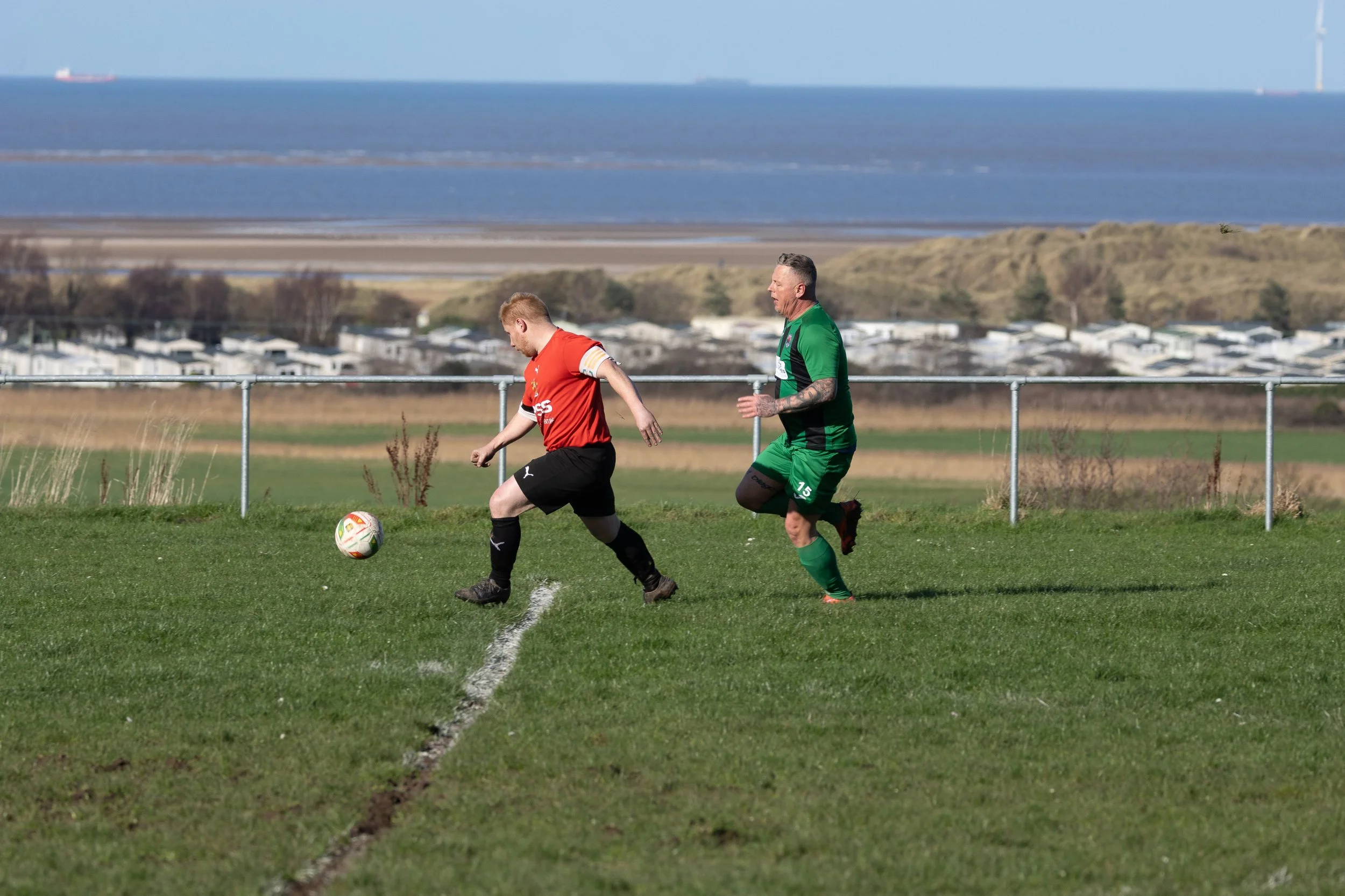 Two soccer players chase after a ball on a grassy field, with a view of water and horizon in the background.