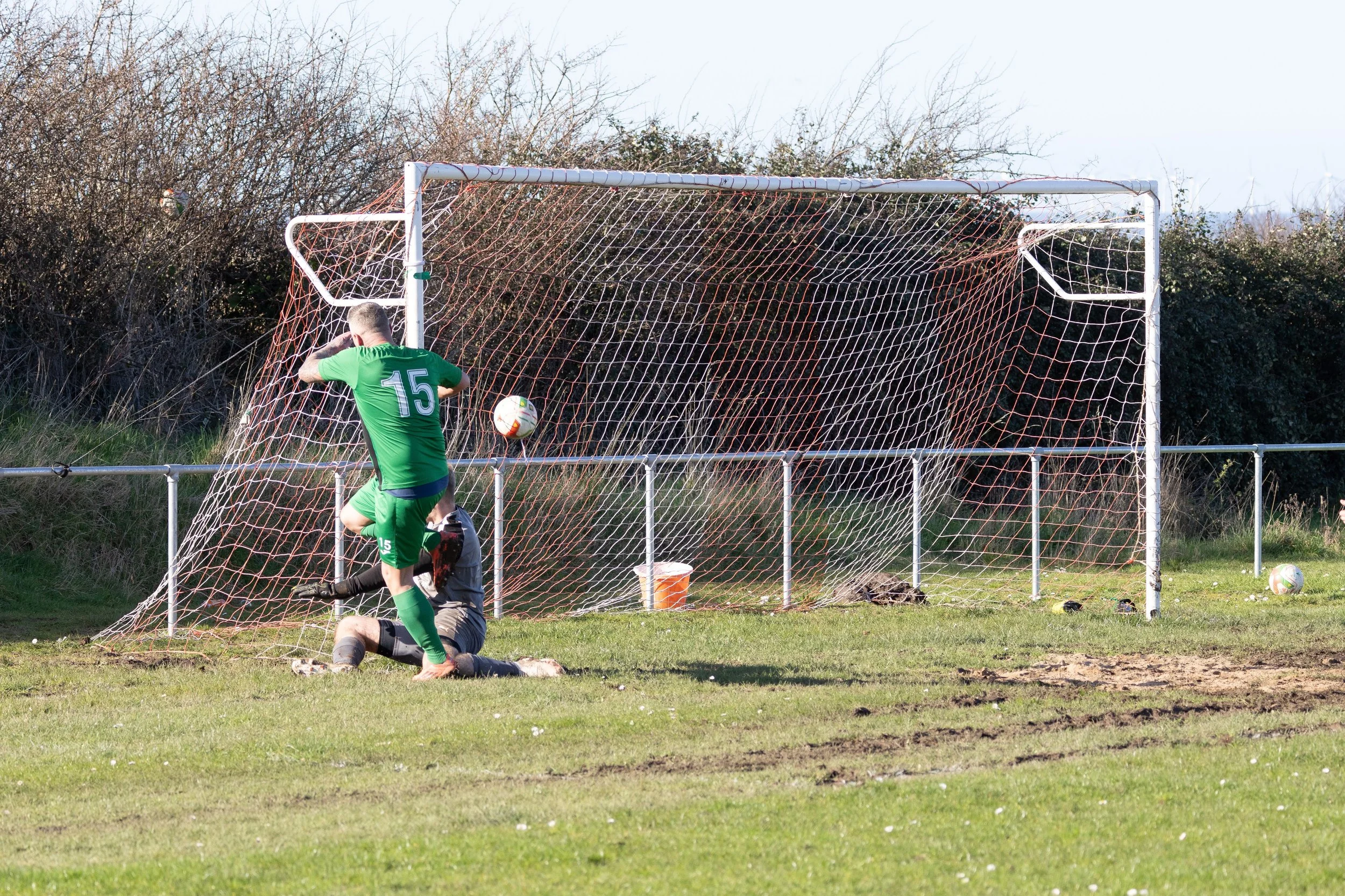 A soccer player in a green jersey and shorts, with the number 15 on his back, is jumping near the goal after a missed shot. The goalkeeper, dressed in black, is sitting on the ground near the goalpost. A soccer ball is in mid-air near the goal, and a