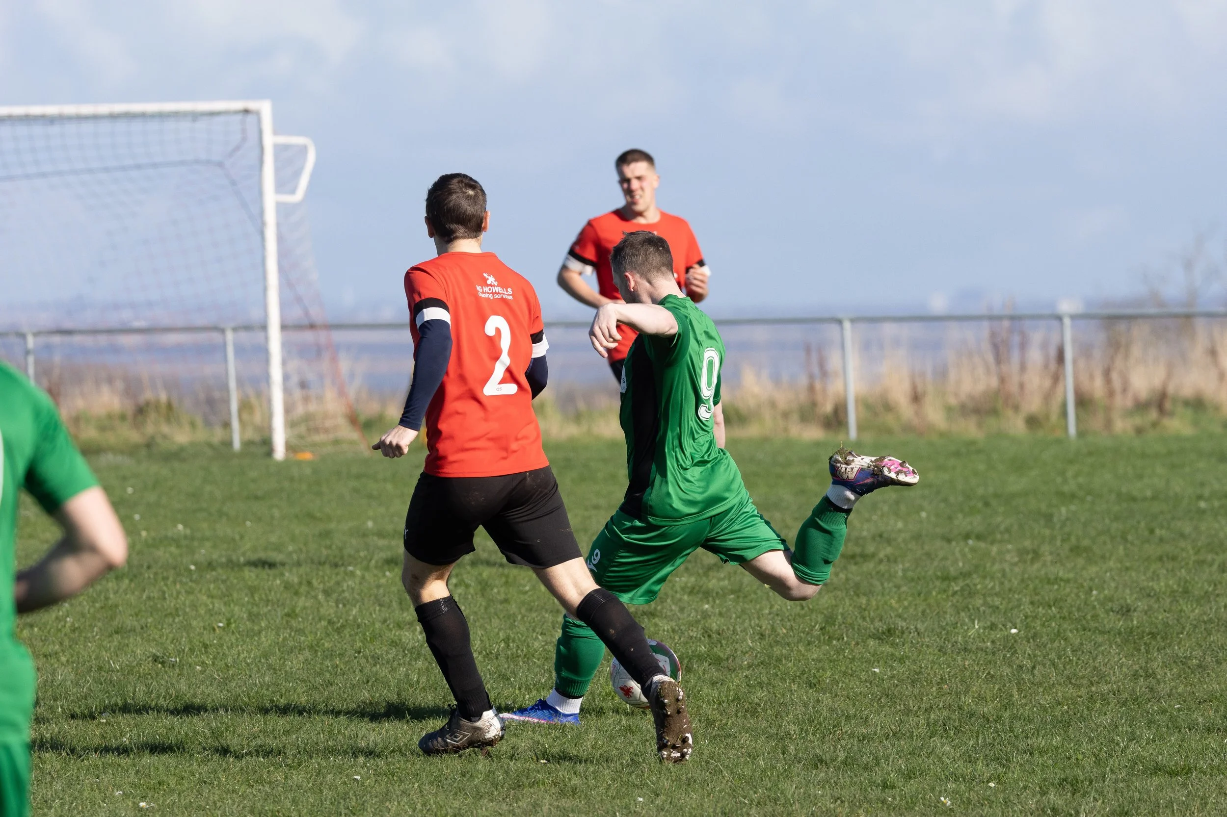 Soccer players in green and red uniforms competing for the ball on a grassy field with goalpost in background.