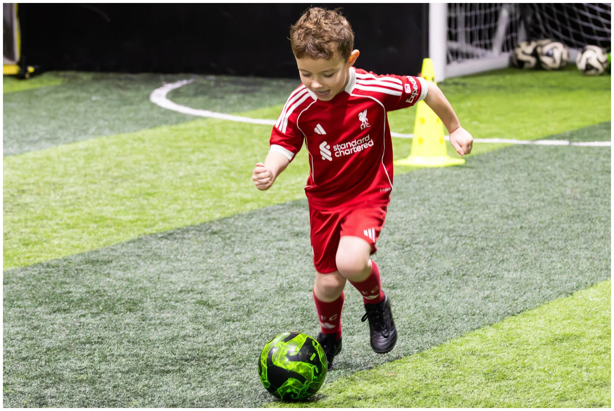 A young boy in a red soccer uniform playing soccer indoors, dribbling a black and green soccer ball.