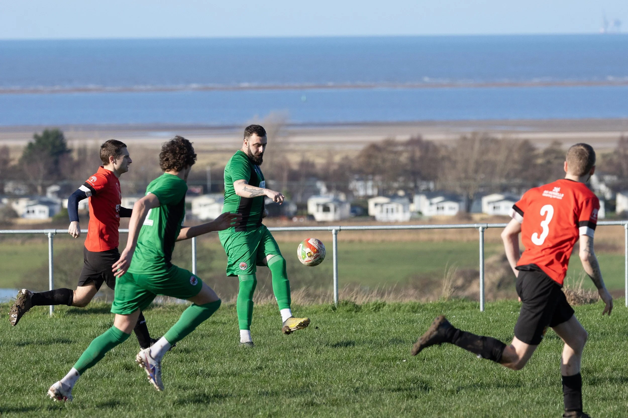Soccer players in red and green uniforms playing on a grassy field with a body of water and houses in the background.