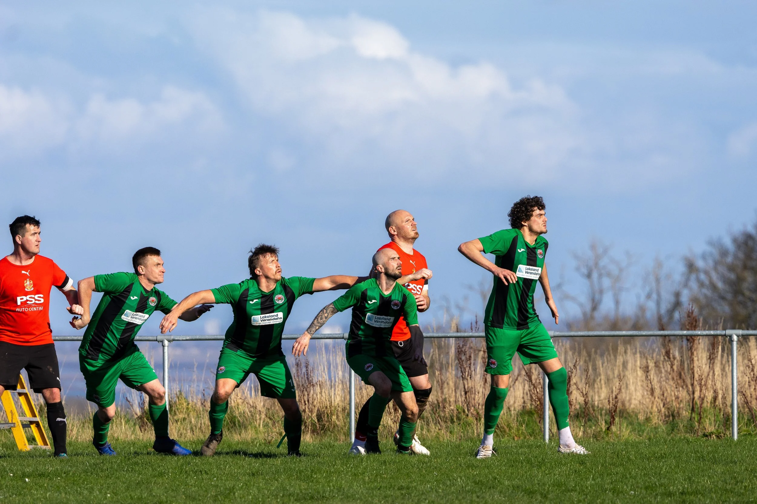 Soccer players in green and red jerseys preparing for a free kick on a grassy field with a blue sky and sparse trees in the background.
