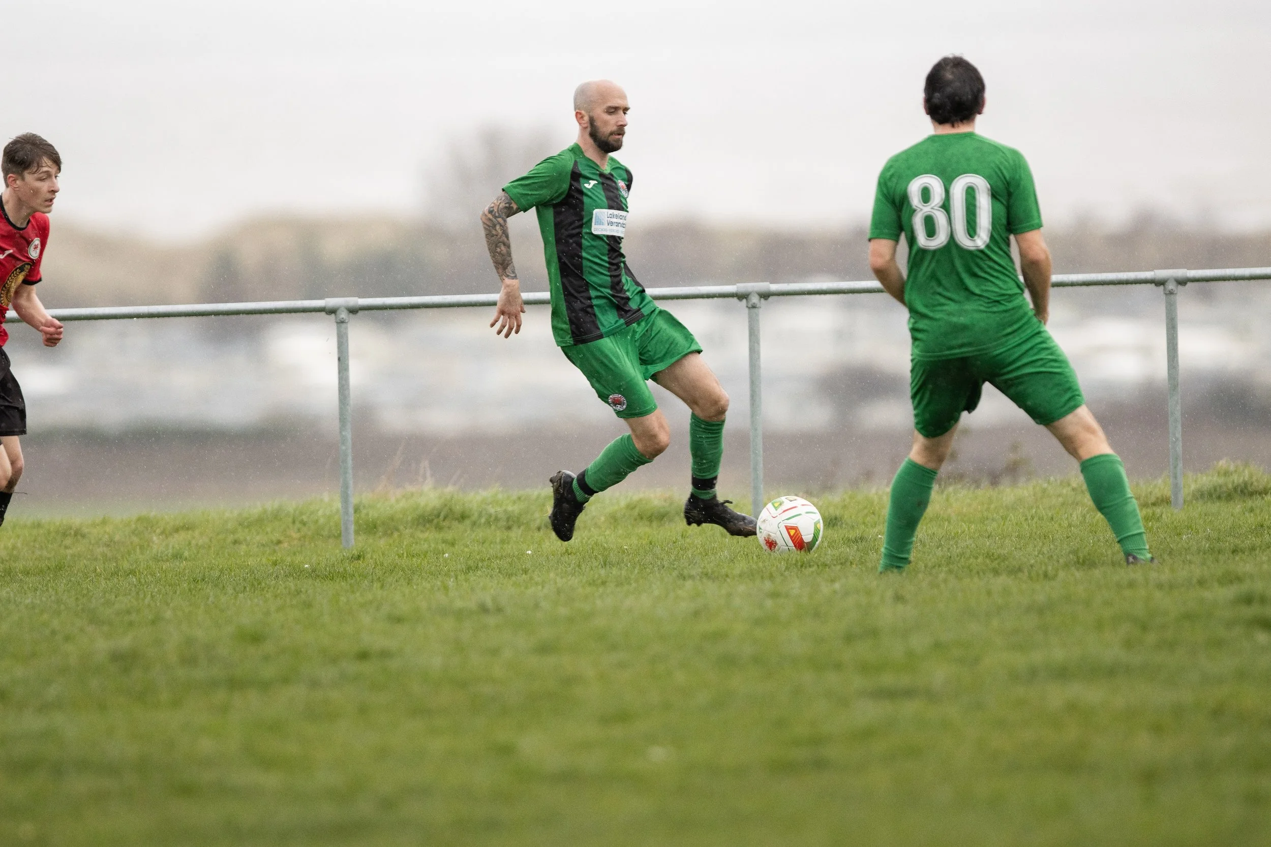 Soccer players on a green field during a game, one in a black and green uniform with a tattooed arm, another in green with the number 80 on the back, and a third in red and black on the left side.