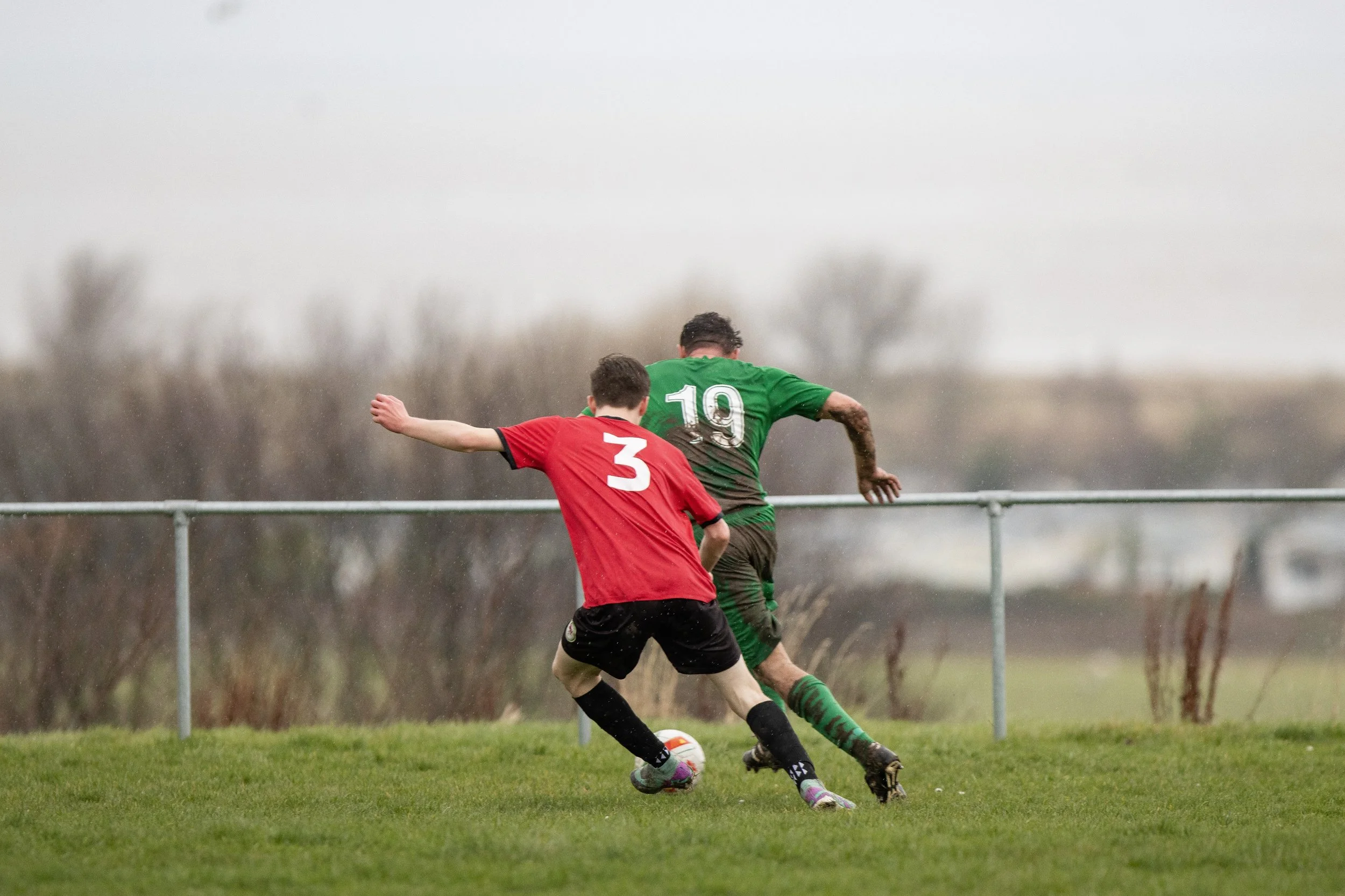 Two soccer players, one in red and black with the number 3 and the other in green with the number 19, compete for the ball on a grassy field with a metal railing and cloudy sky in the background.