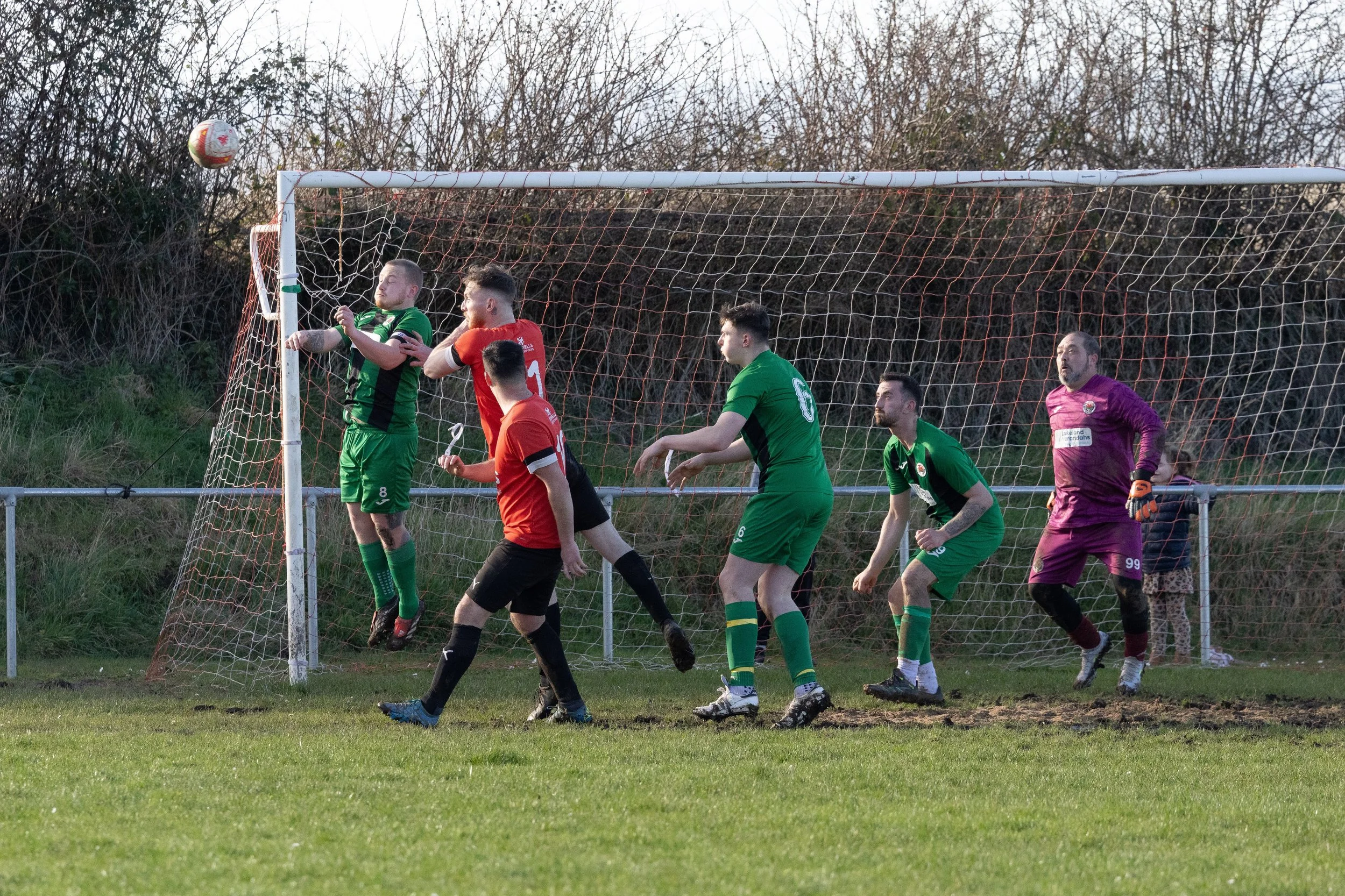 Soccer players in green and red uniforms trying to head a ball in front of the goal, goalkeeper in purple preparing to save, during a match on a grassy field.