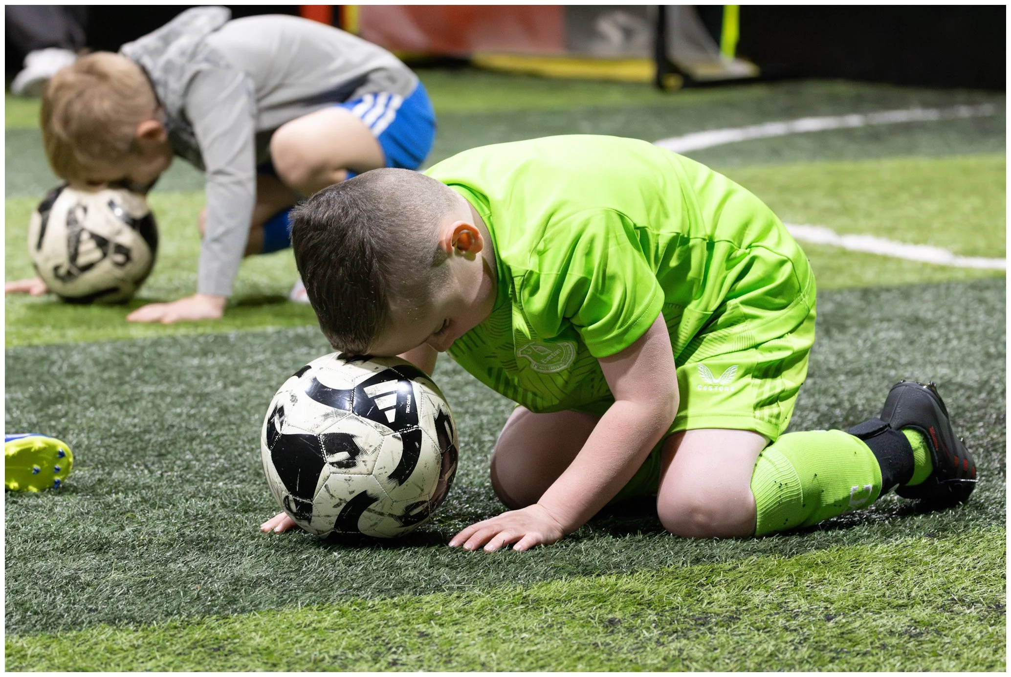 Children kneeling on a soccer field with their foreheads and hands touching the ground, each resting their heads on a soccer ball, participating in a moment of prayer or reflection during a youth soccer practice or game.