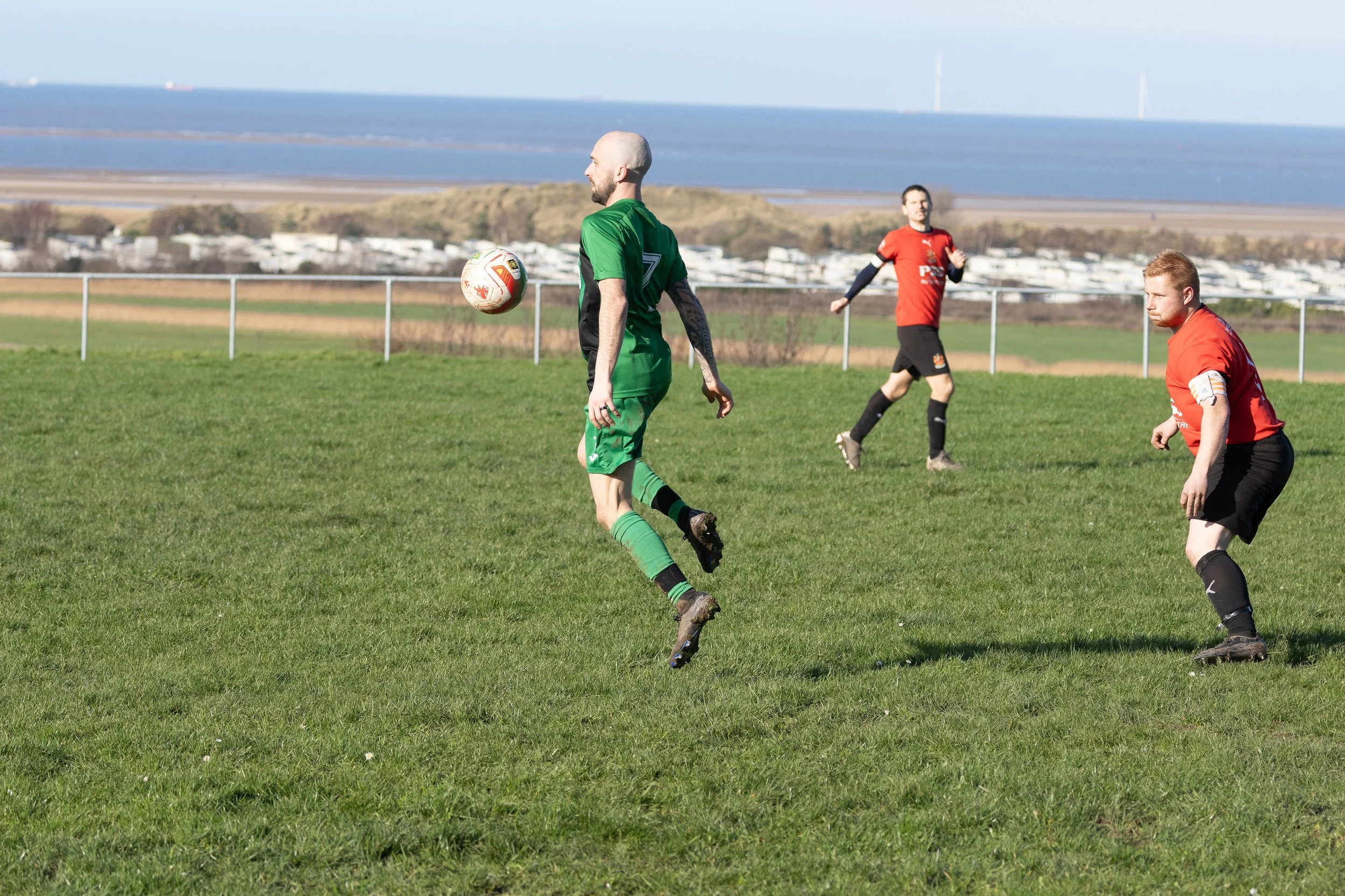 Soccer players on a field, one in green uniform heading the ball, two in red uniforms nearby, with a fence, countryside, and water in the background.