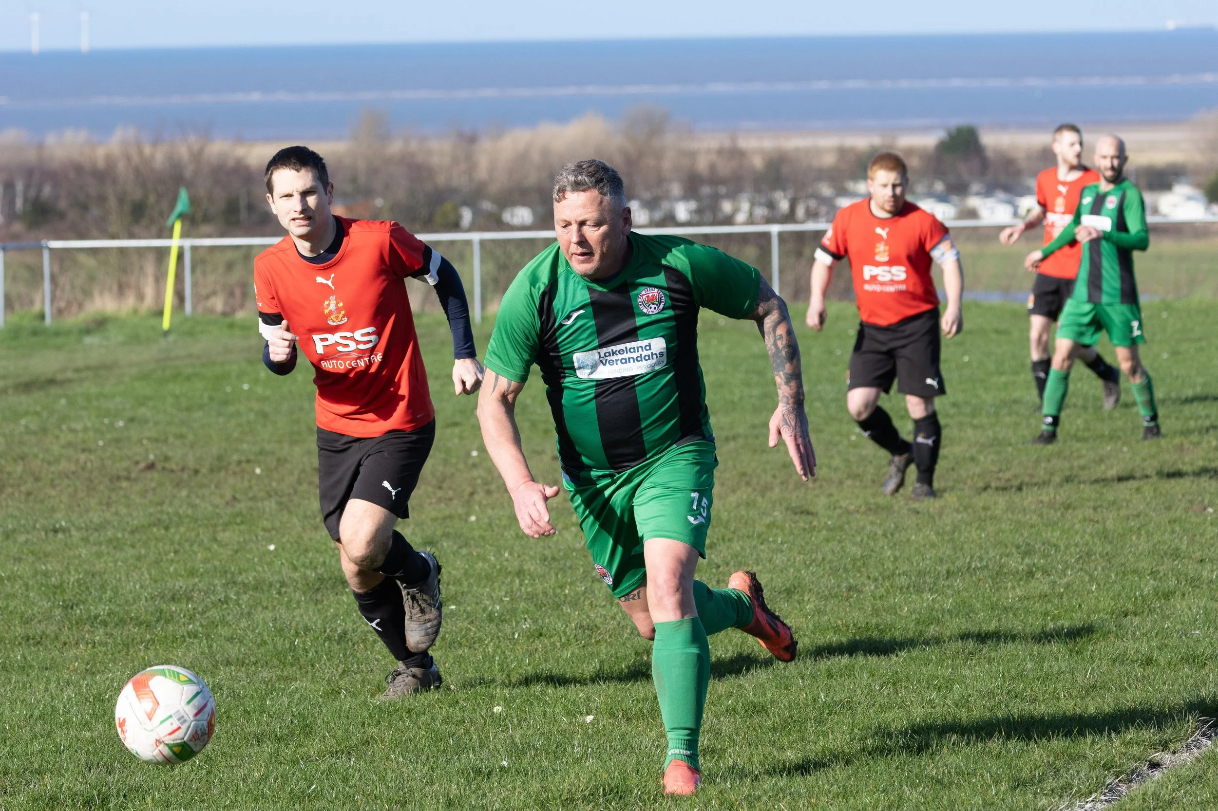 Soccer players on a grassy field during a match, with one player in a green and black uniform approaching the ball and others in red and black nearby.