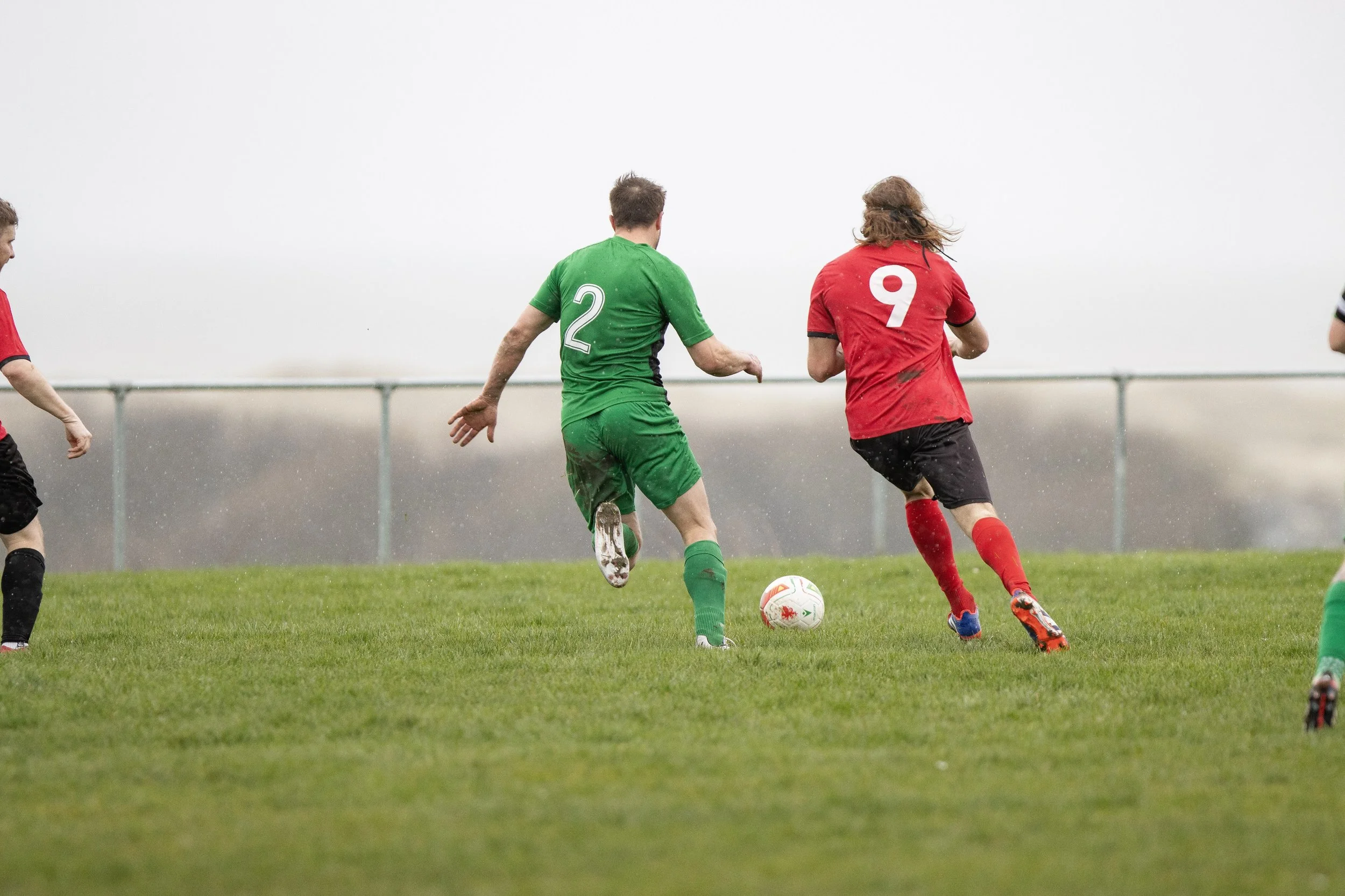 Soccer players in red and green jerseys chasing a ball on a grassy field, with a blurred background and overcast sky.
