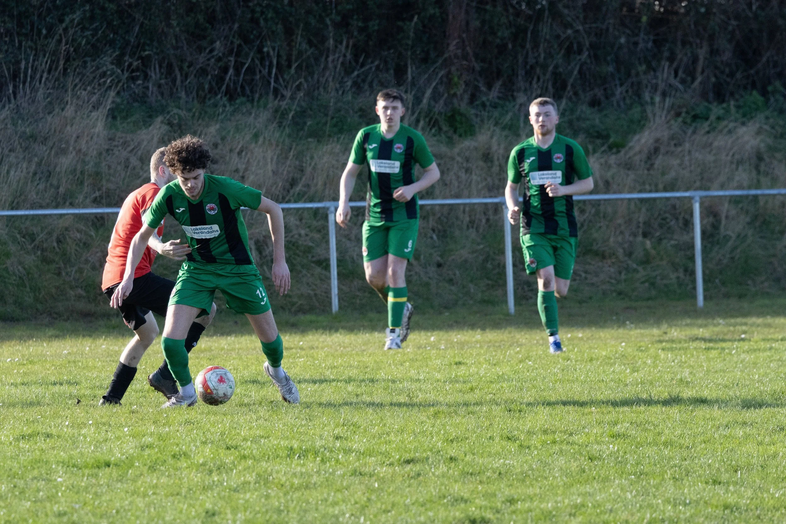 Four young male soccer players, three in green uniforms and one in an orange and black uniform, playing on a grassy field with a background of trees and a metal fence. One player in green is dribbling the ball while the others are running.