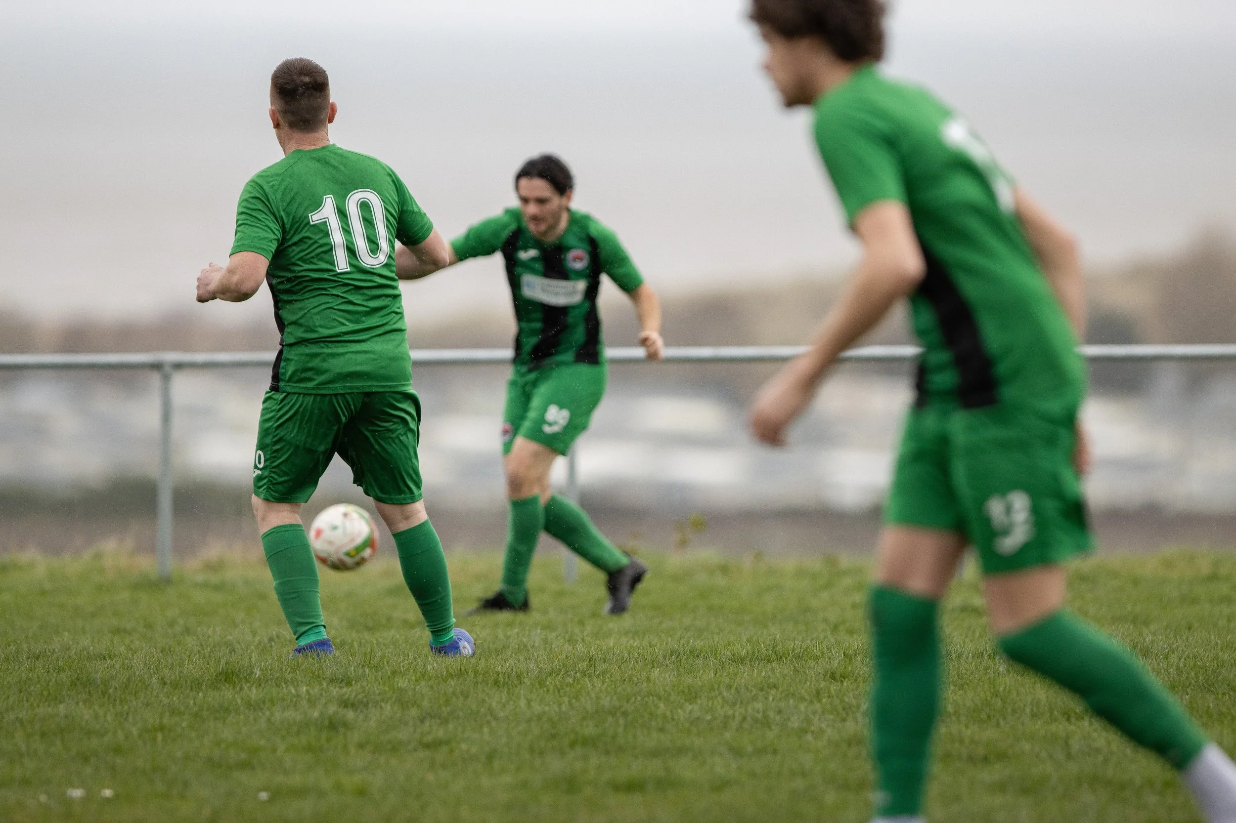 Three soccer players in green uniforms on a grass field, with one player kicking a ball while the others are nearby, and a cloudy sky in the background.