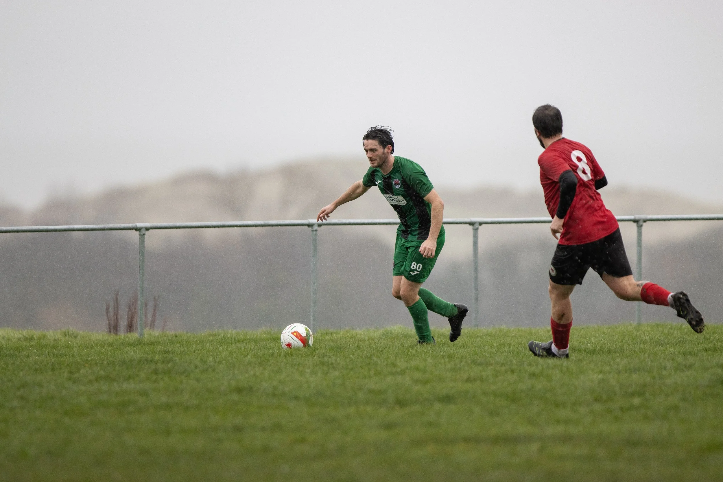 Soccer player in green uniform dribbling ball while facing defender in red uniform on an outdoor field.