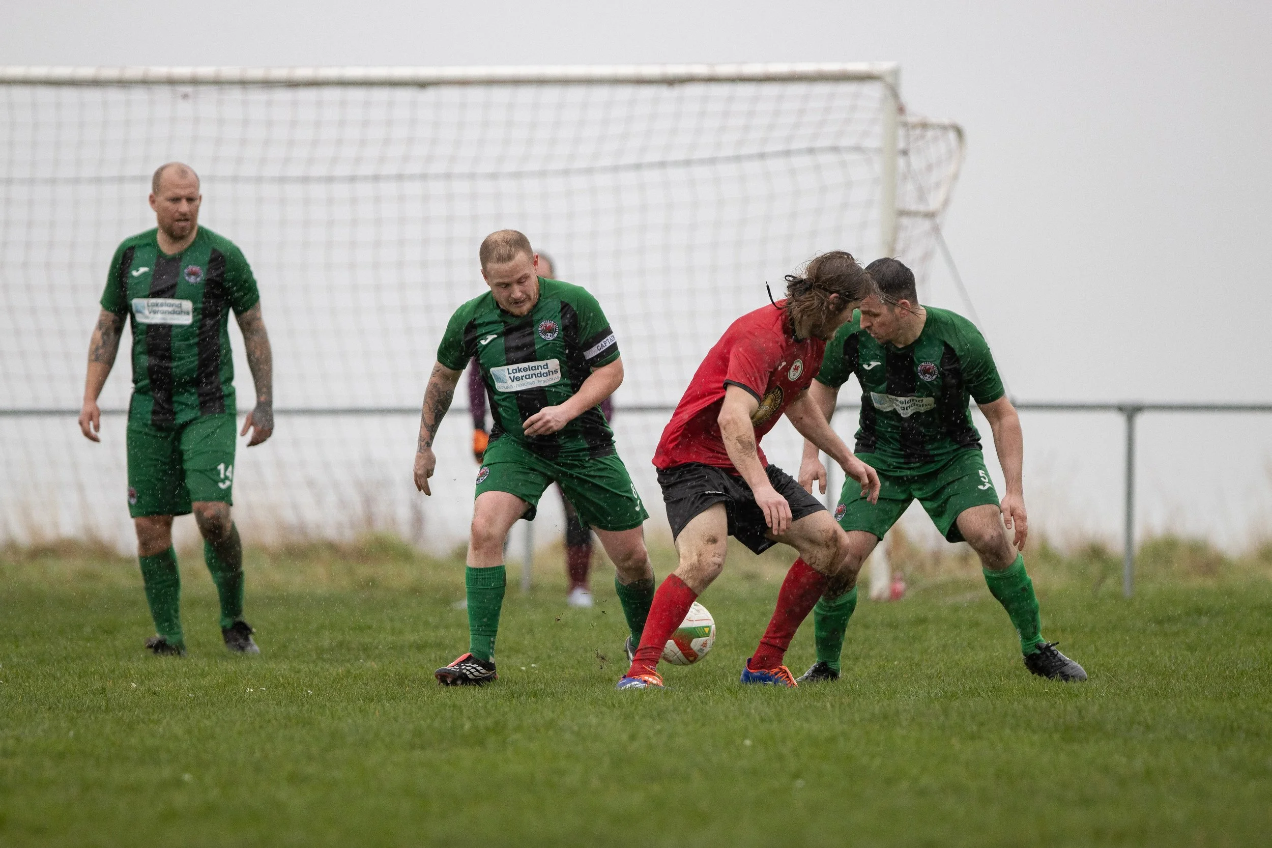 Soccer players in green and red jerseys competing for the ball on a rainy day.