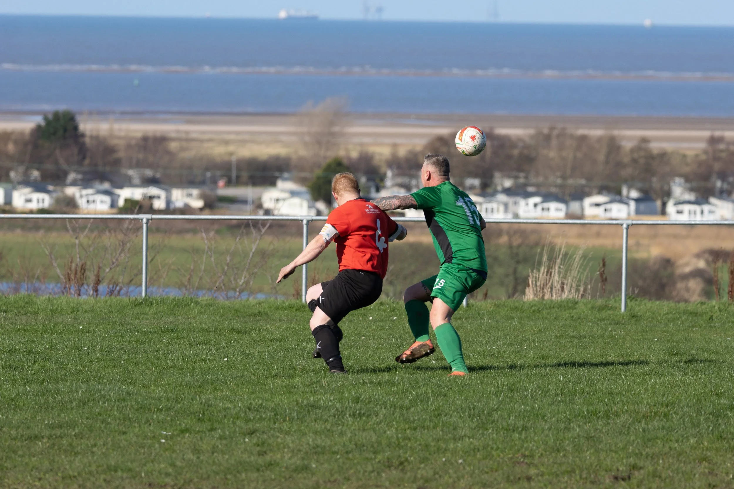 Two men playing soccer on a grassy field, with a body of water and buildings in the background.