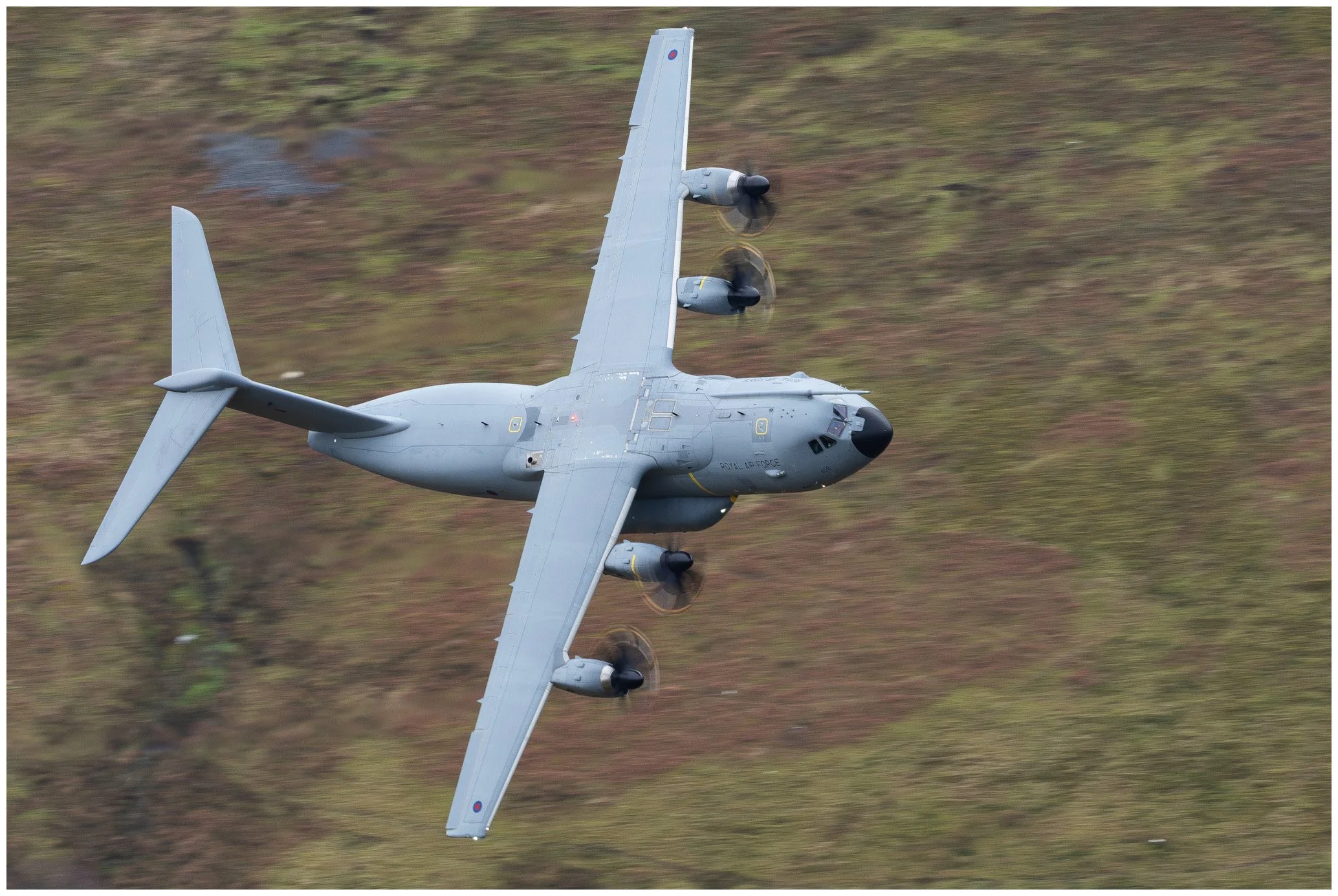 A grey military aircraft flying at an angle over a grassy landscape with blurred background.