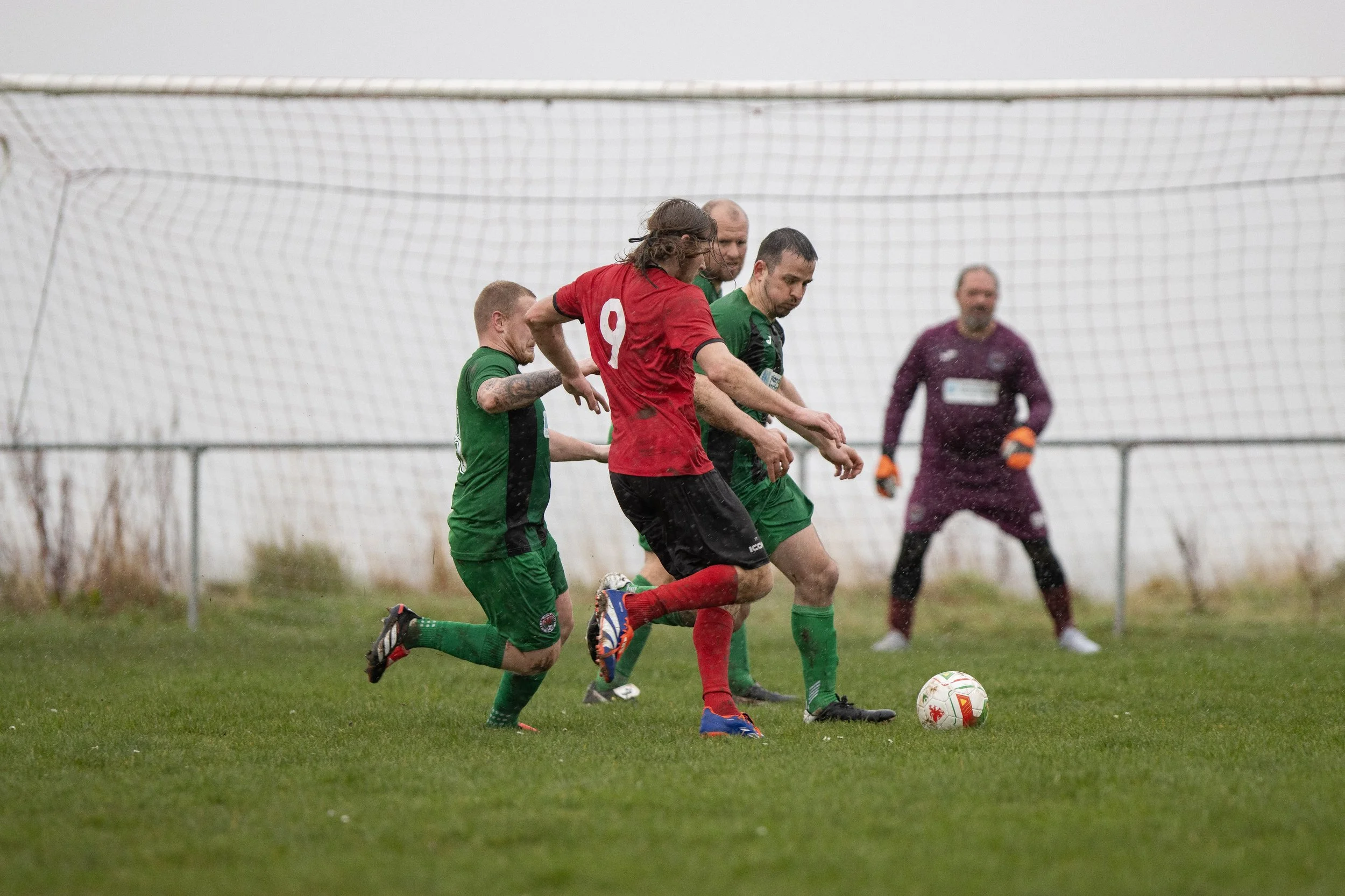 Soccer players competing for the ball on rainy day with a goalie in the background.