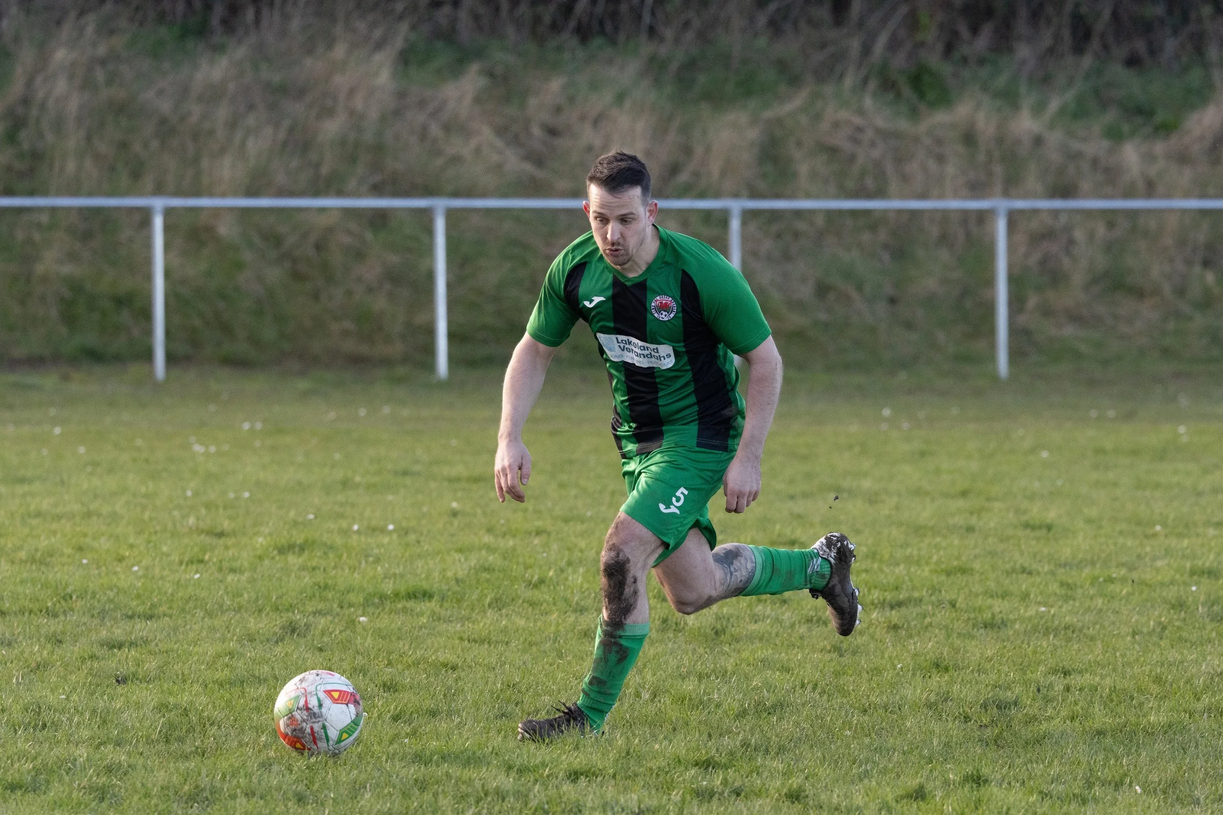 A male soccer player in a green and black uniform is on a grassy field, preparing to kick a soccer ball. He has short dark hair and some mud on his right knee and left shin.