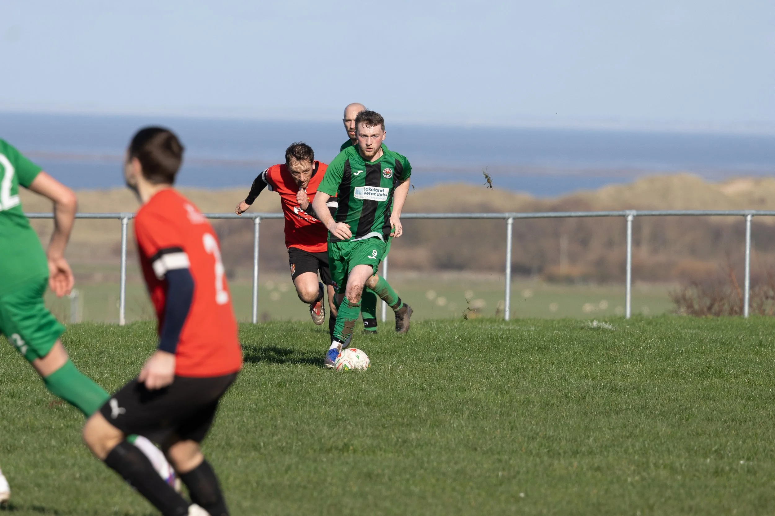 Soccer game with players in green and red jerseys on a grassy field near the coast, with a fence and hills in the background.