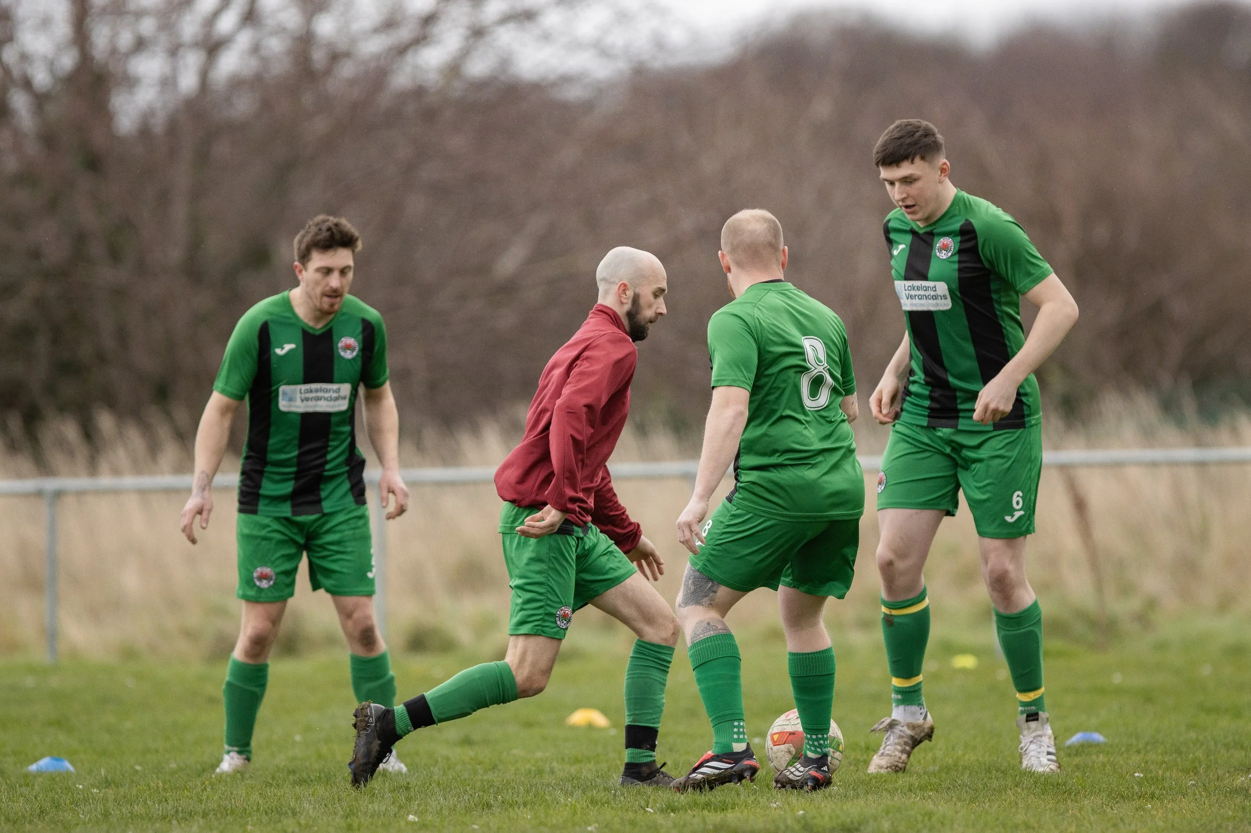 Four soccer players on a field, three in green jerseys and one in a red jacket, appear to be in a discussion over a soccer ball during practice or a game.