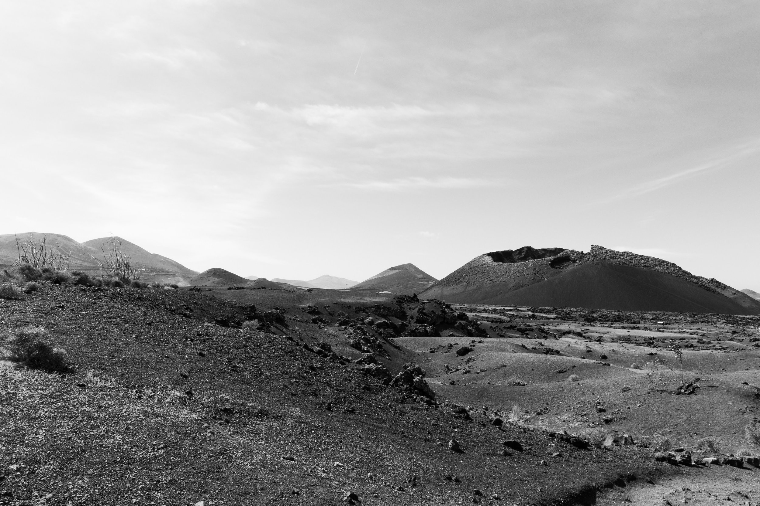 Black and white photo of a barren, mountainous desert landscape with rough terrain, sparse vegetation, and hills in the distance under a cloudy sky.