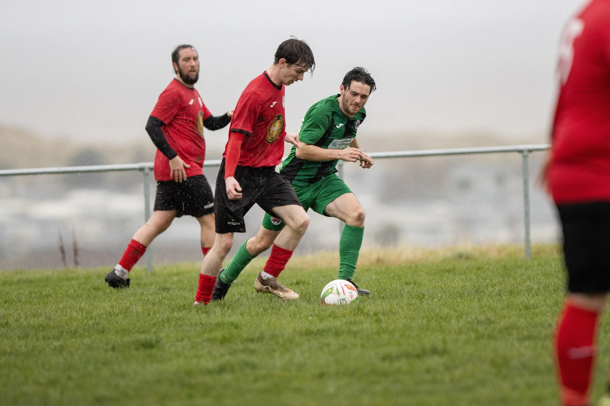 Soccer players battling for the ball during a match in rainy weather, with a foggy background and green grass field.