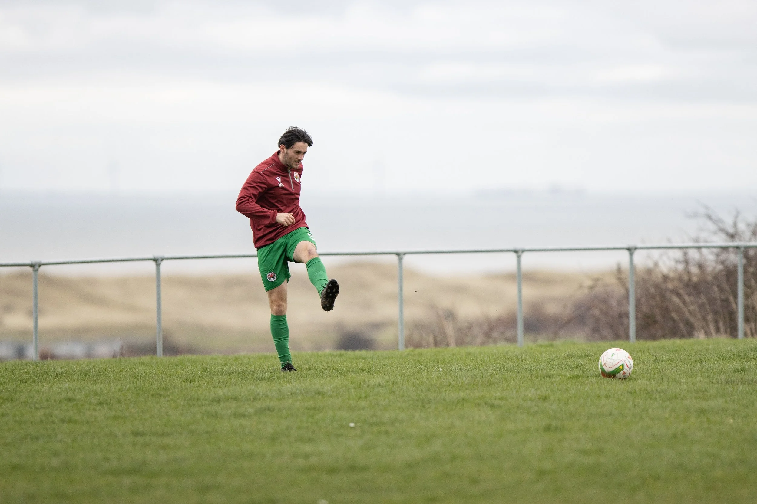 A man in a maroon jacket, green shorts, and green socks is kicking a soccer ball on a grassy field near a fence, under a cloudy sky.