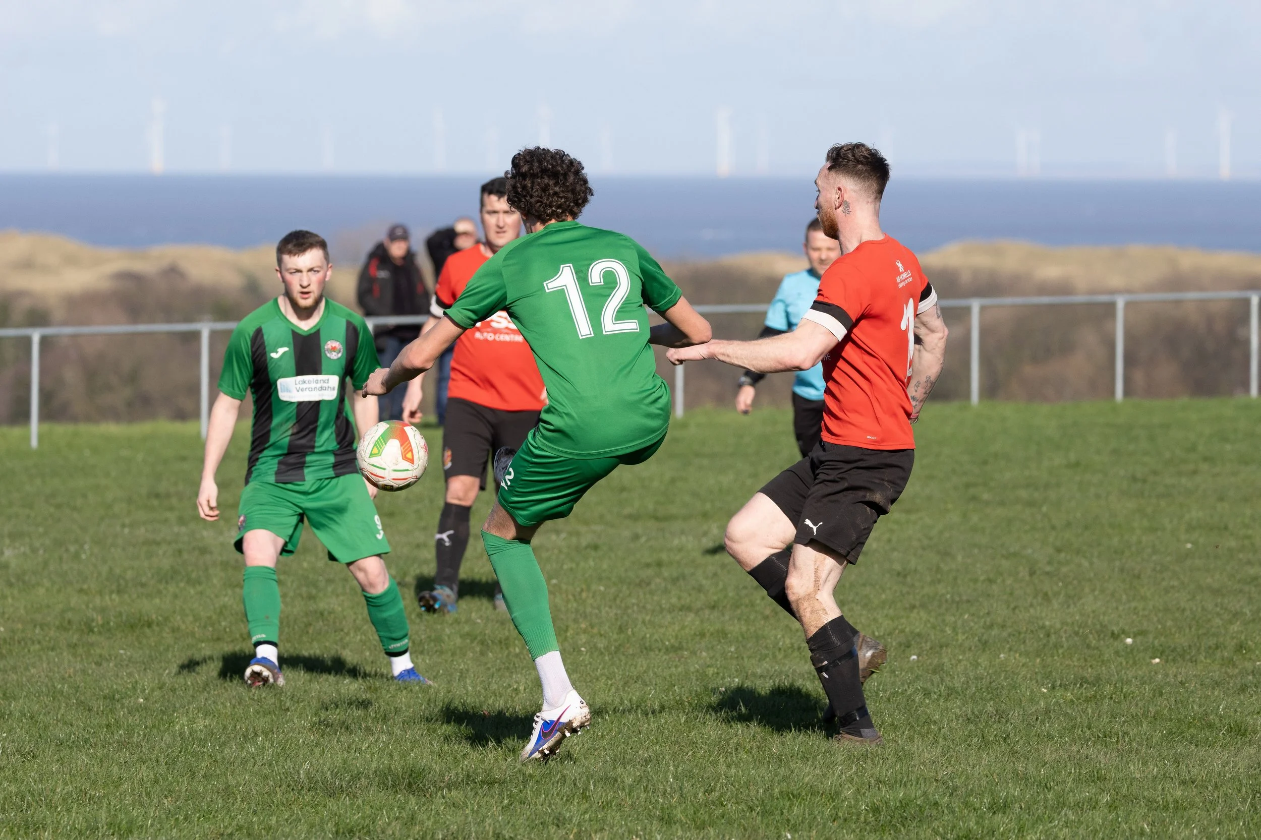 Soccer match with players in green and red jerseys on a grassy field, with a blue sky and wind turbines in the background.