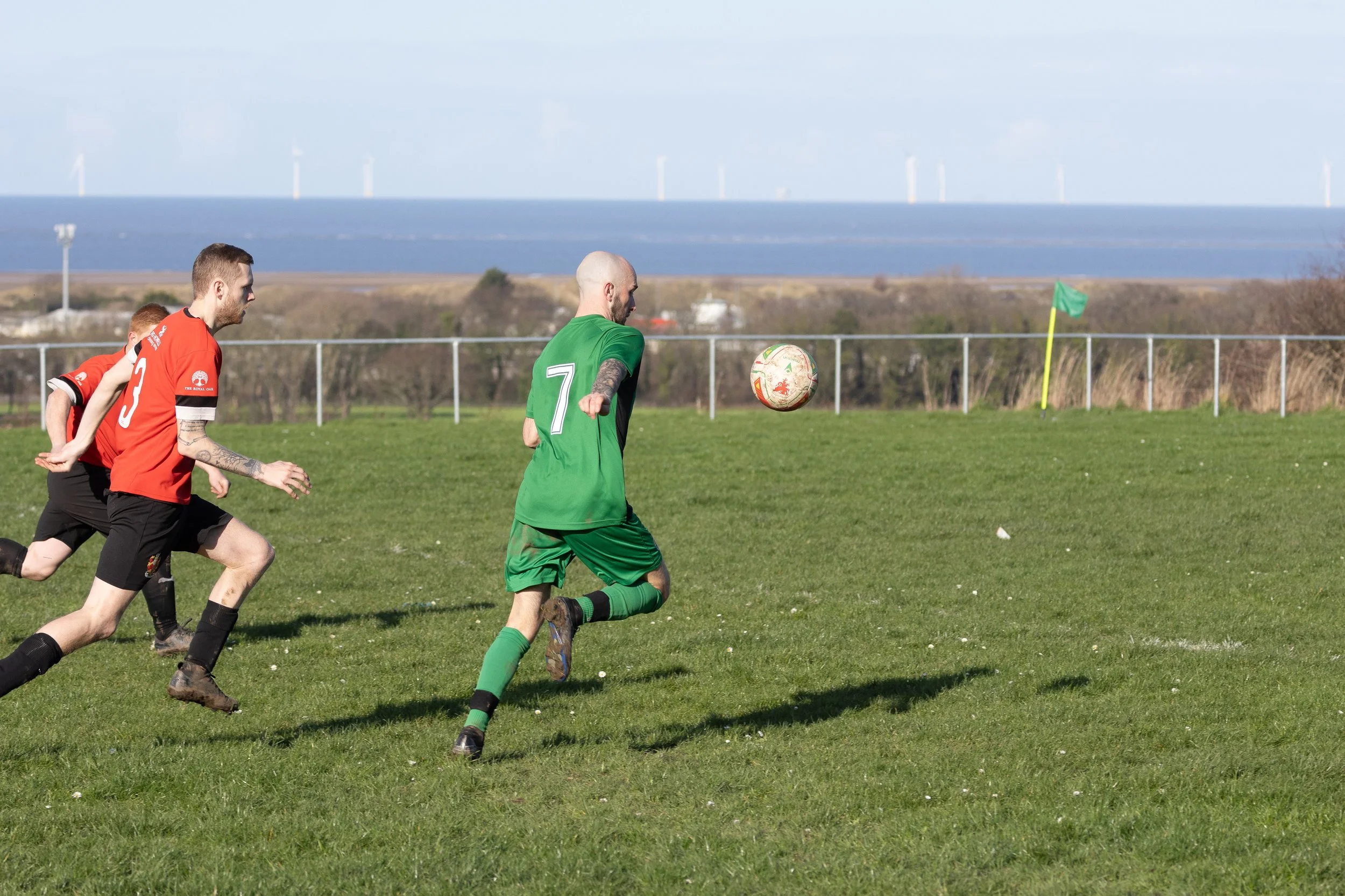 Soccer players in red and green jerseys chase the ball on a grassy field near the coast with wind turbines in the background.