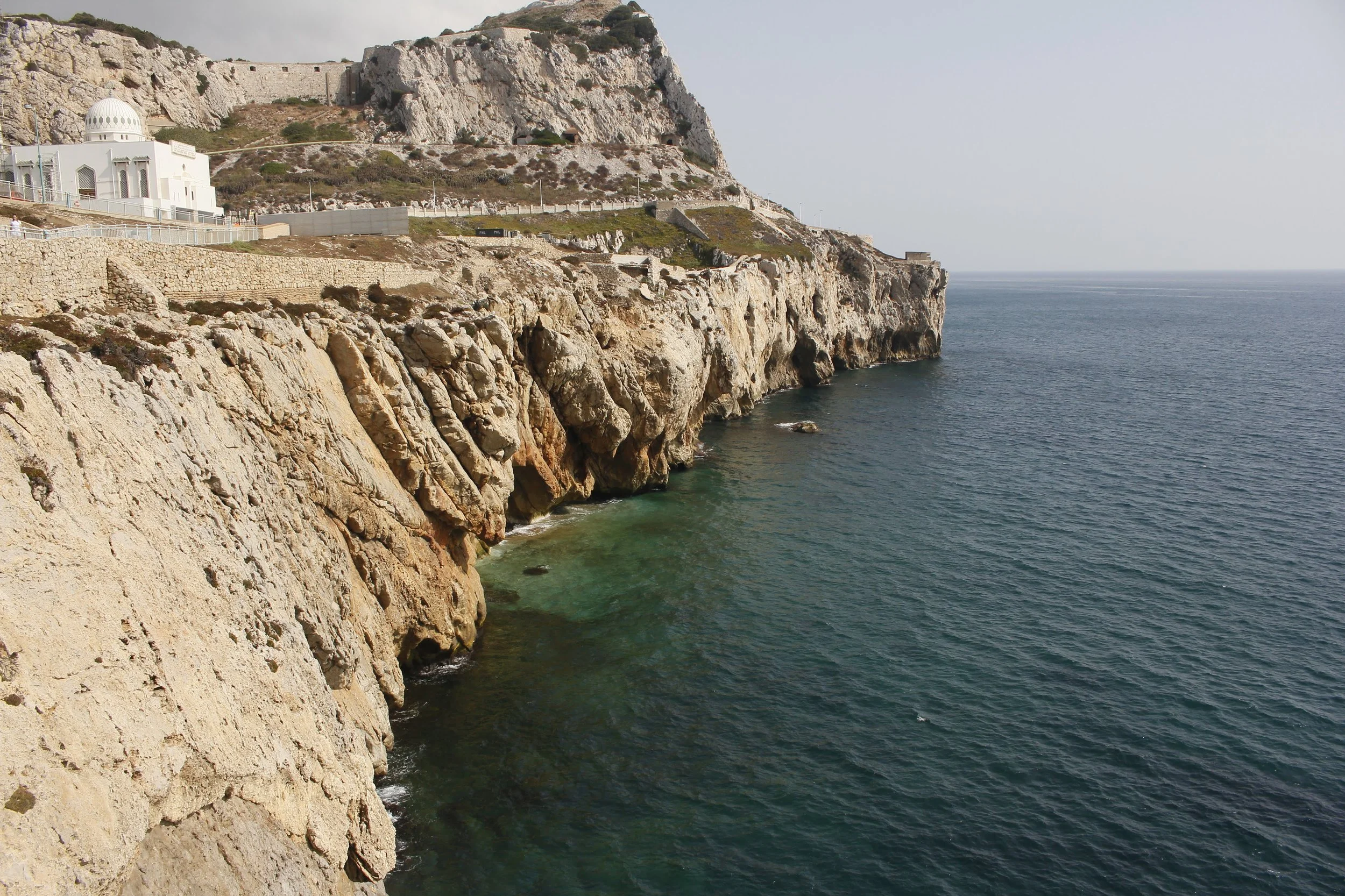 Cliffs overlooking the ocean with a small white building and rocky shoreline in the distance.