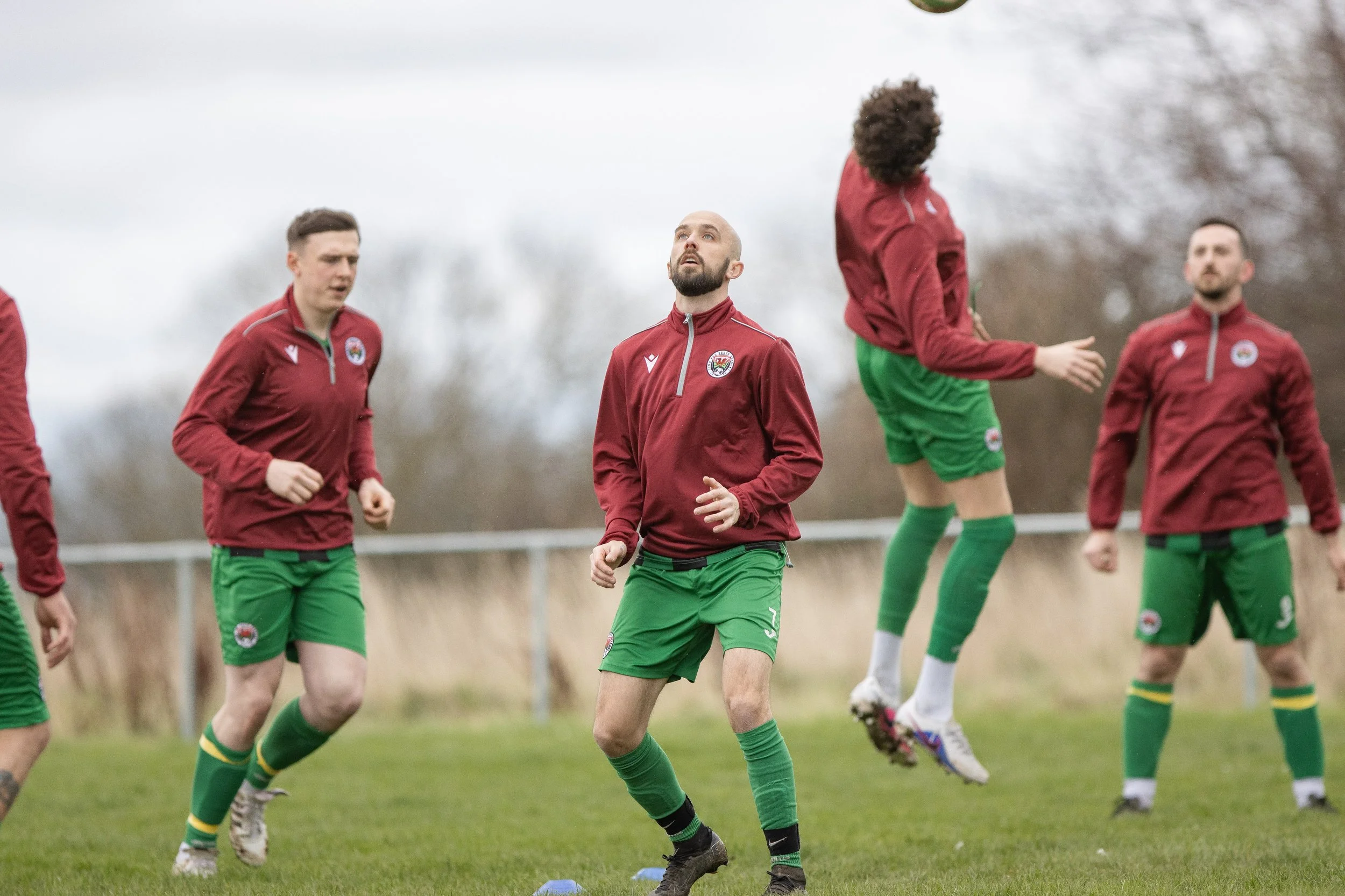 Group of soccer players in maroon jerseys and green shorts practicing on a grass field during the daytime.