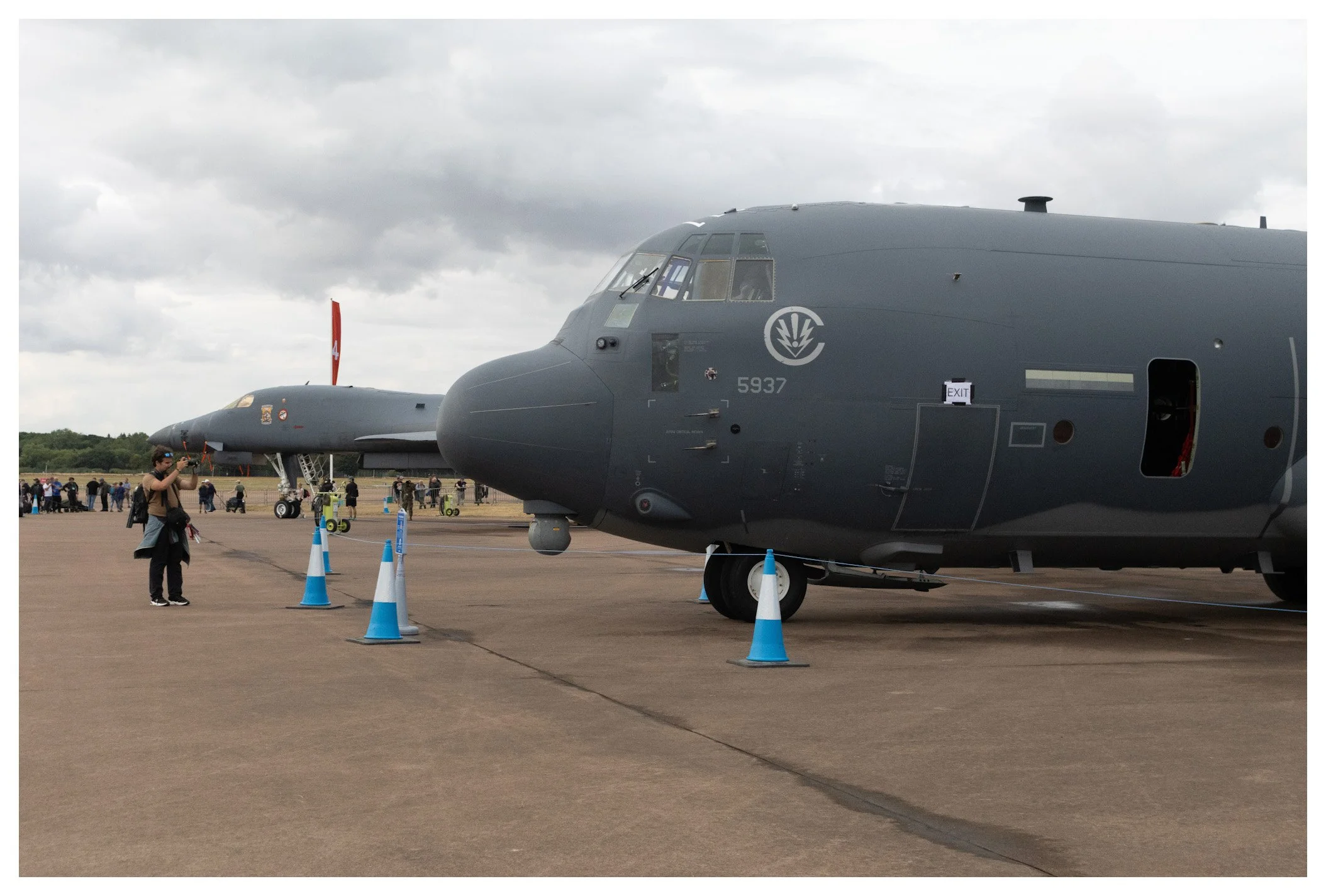 Two large military aircraft on display on an airfield, with people and children observing and taking photos. The foreground features part of a dark gray aircraft with a large nose and cockpit, and the background shows another aircraft with a red tail
