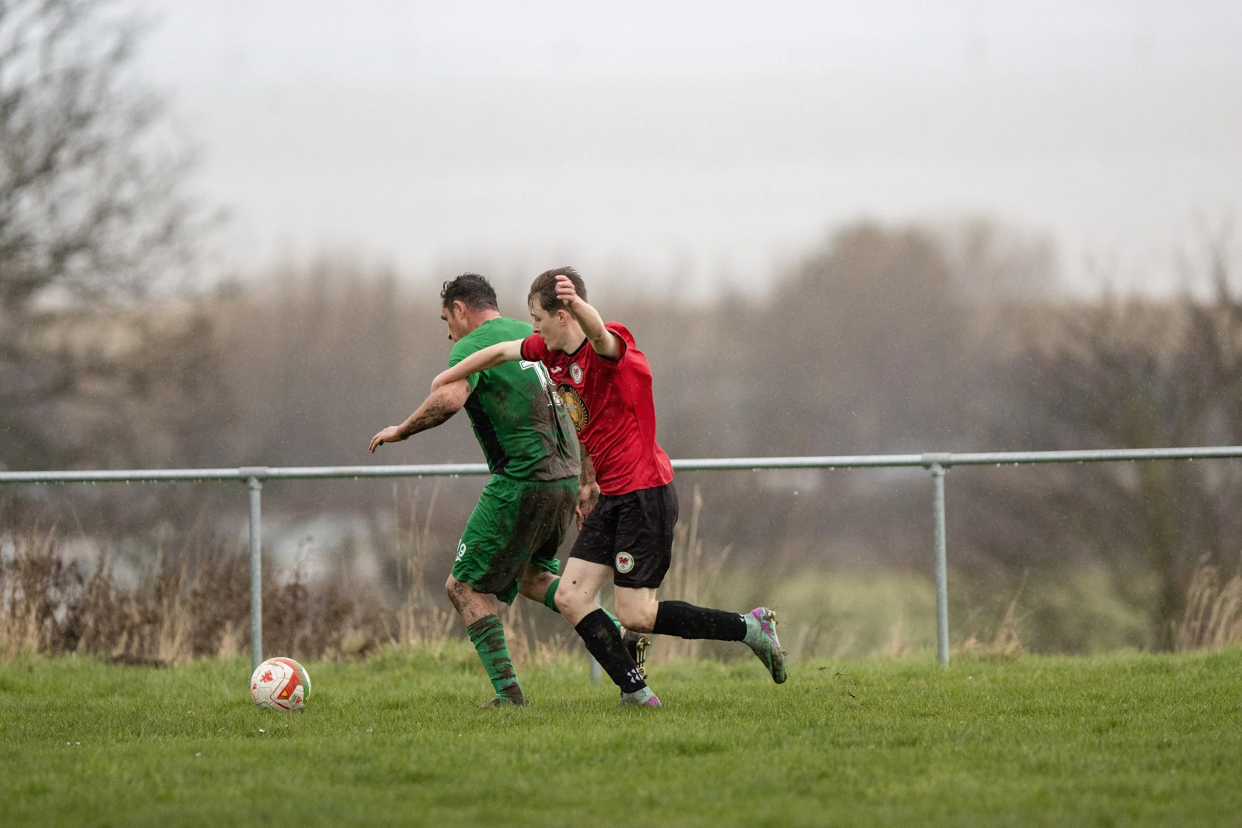 Two soccer players competing for the ball on a muddy field, with a cloudy sky and trees in the background.