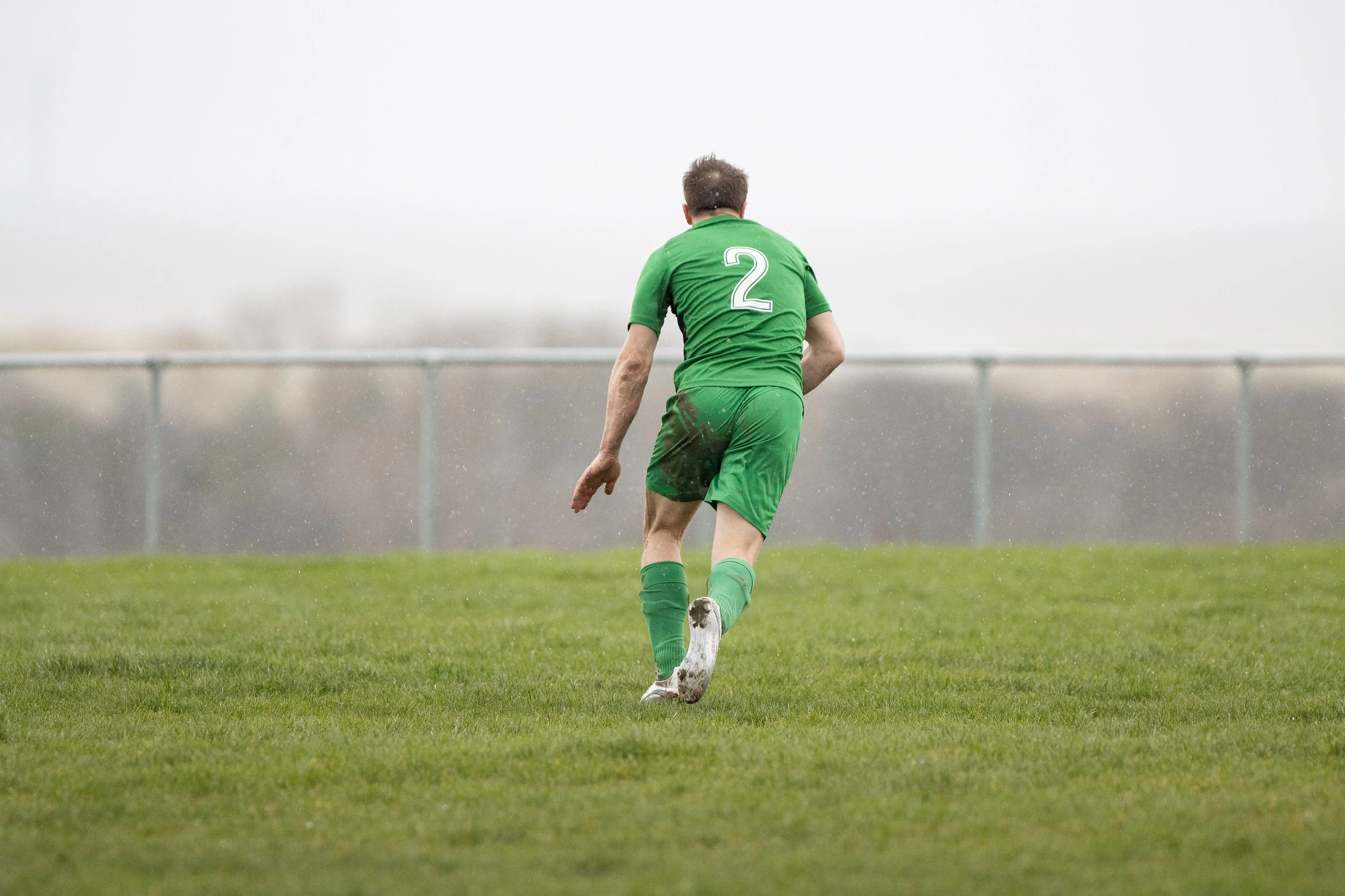 A soccer player in a green uniform with the number 2 on the back, walking on a wet grass field during a game or practice, with a foggy or overcast sky in the background.