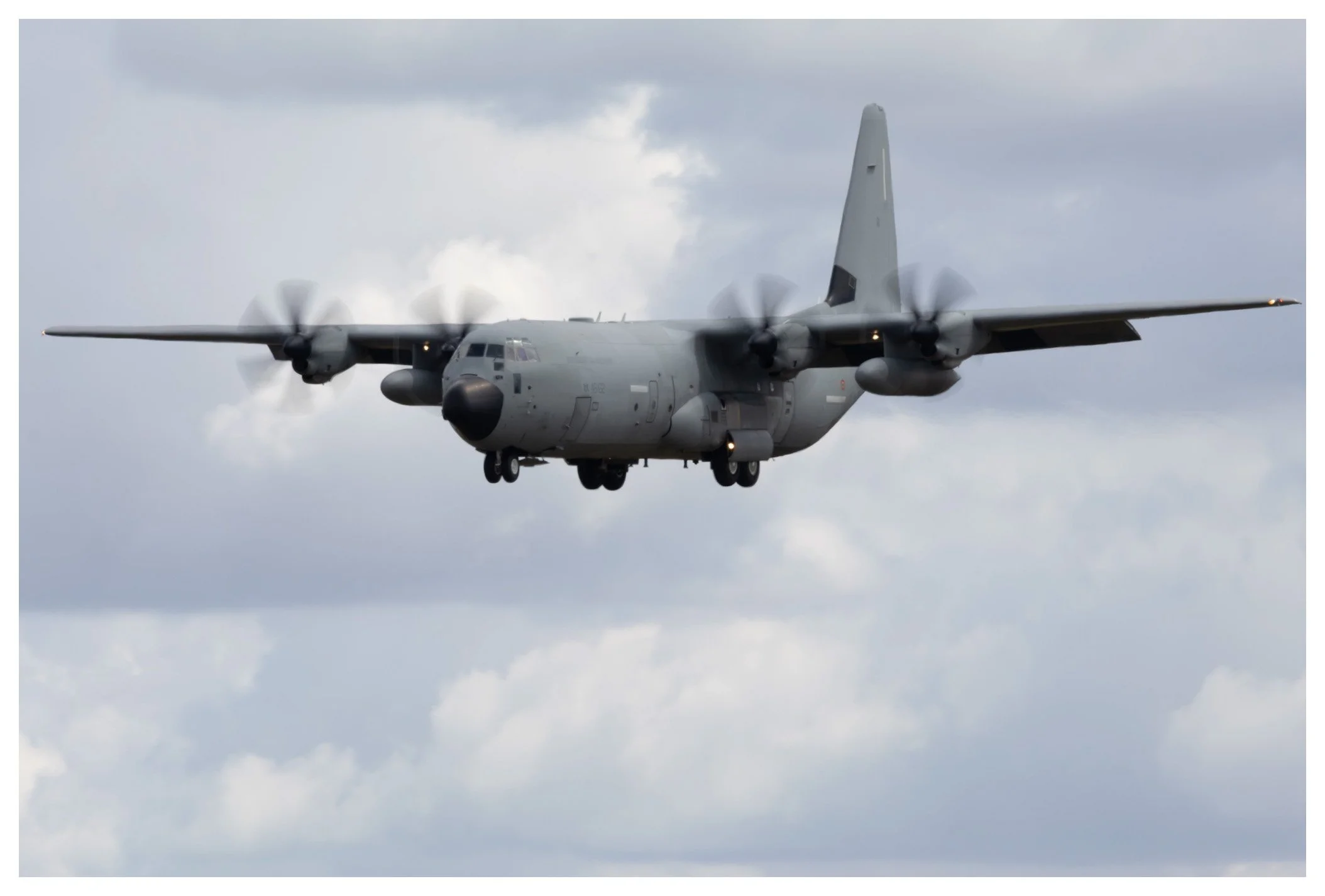 A military cargo plane flying in the cloudy sky with four turbo-prop engines turning.