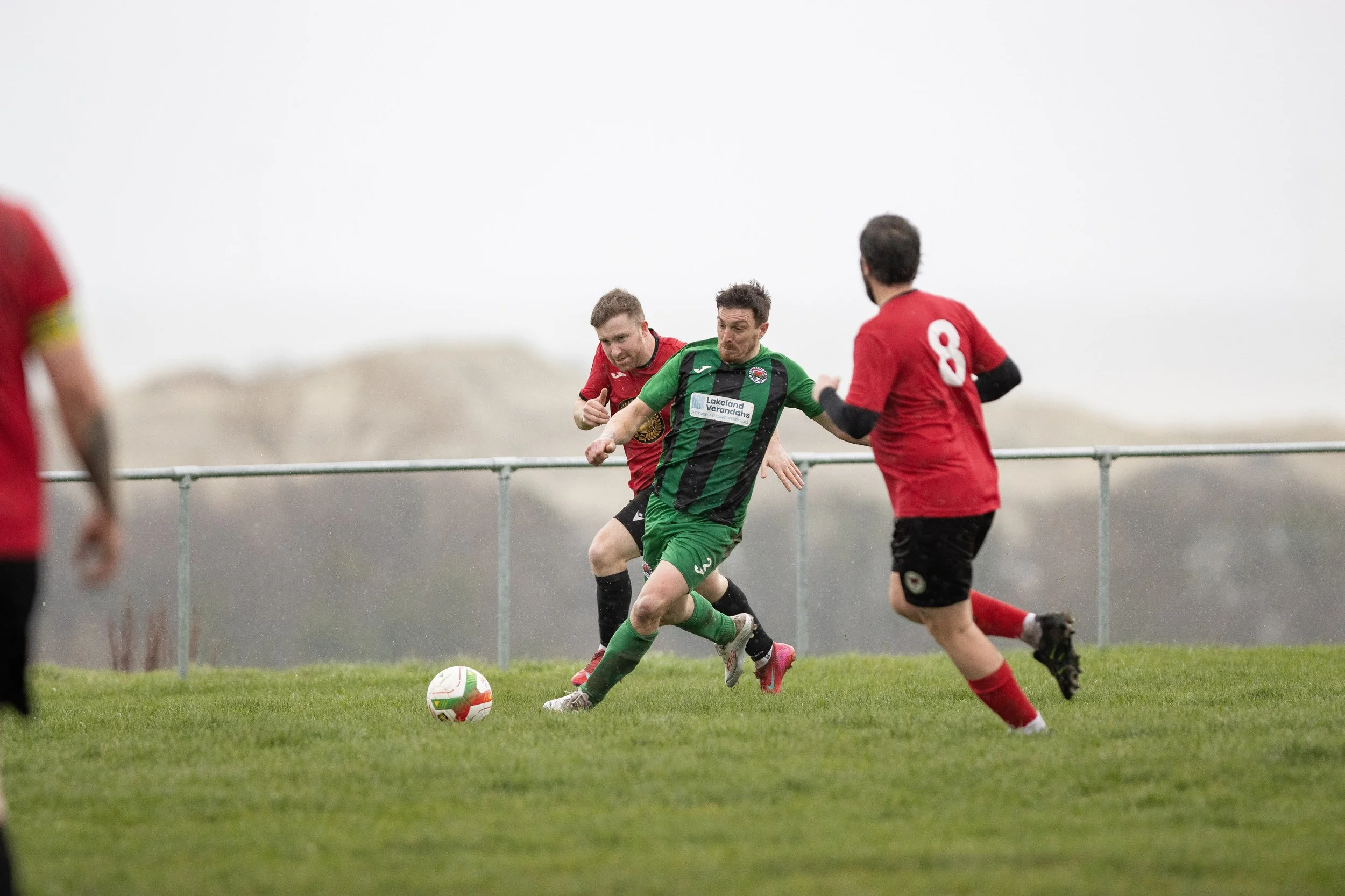 Soccer players in green and red jerseys competing for the ball on a grassy field, with a cloudy sky in the background.