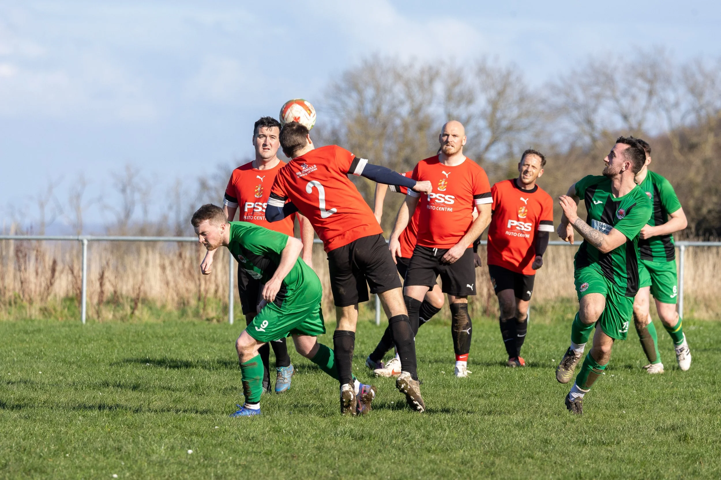 Soccer match with players in red and green uniforms contesting an airborne ball on a grassy field with a fence and trees in the background.