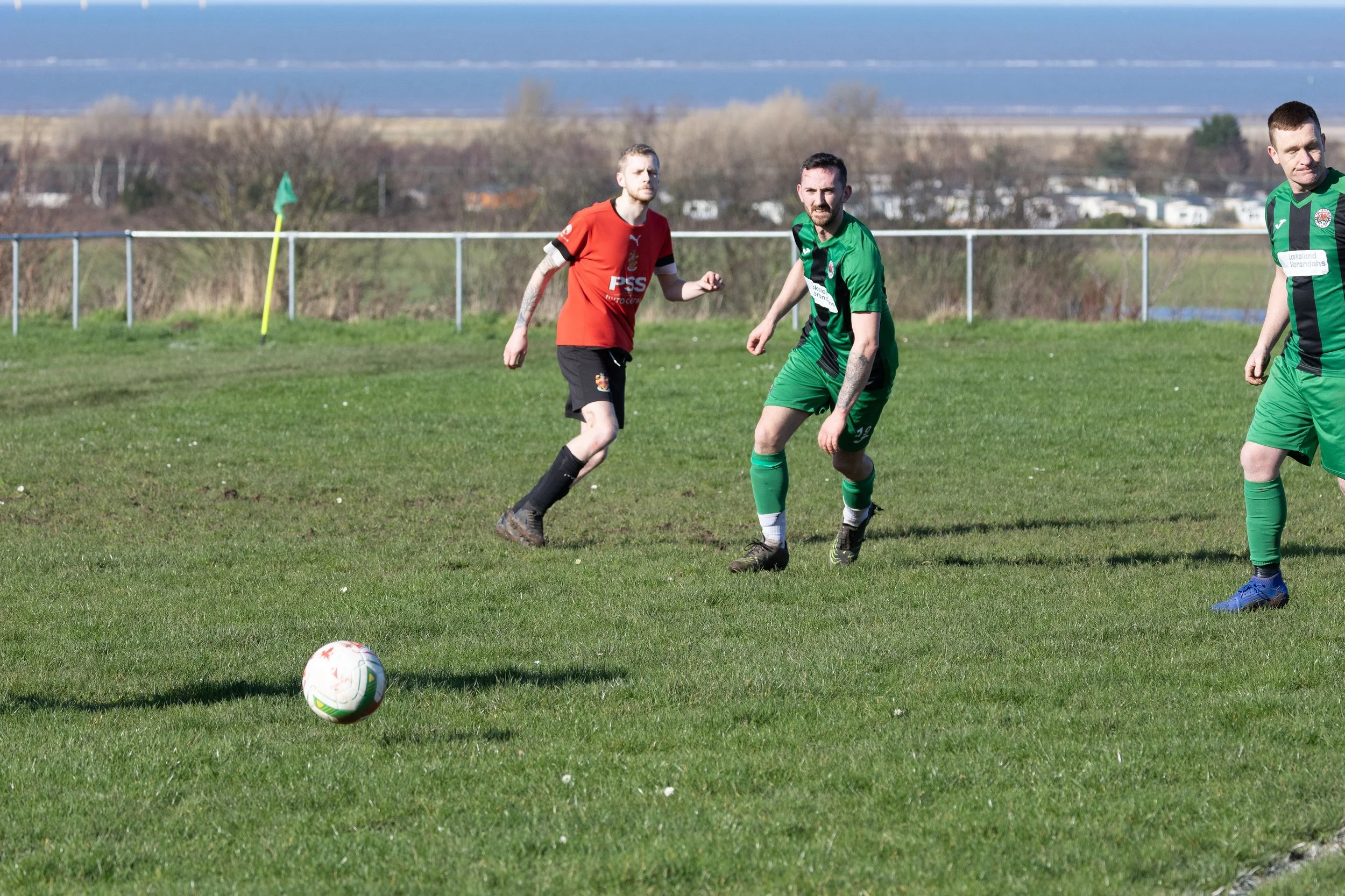 Soccer players in green and red jerseys on a grassy field near a fence, with a scenic view of water and land in the background.