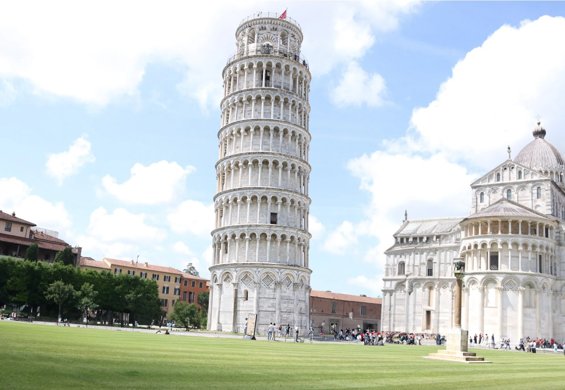 The Leaning Tower of Pisa beside a cathedral with people walking on a grassy area and a partly cloudy sky in the background.