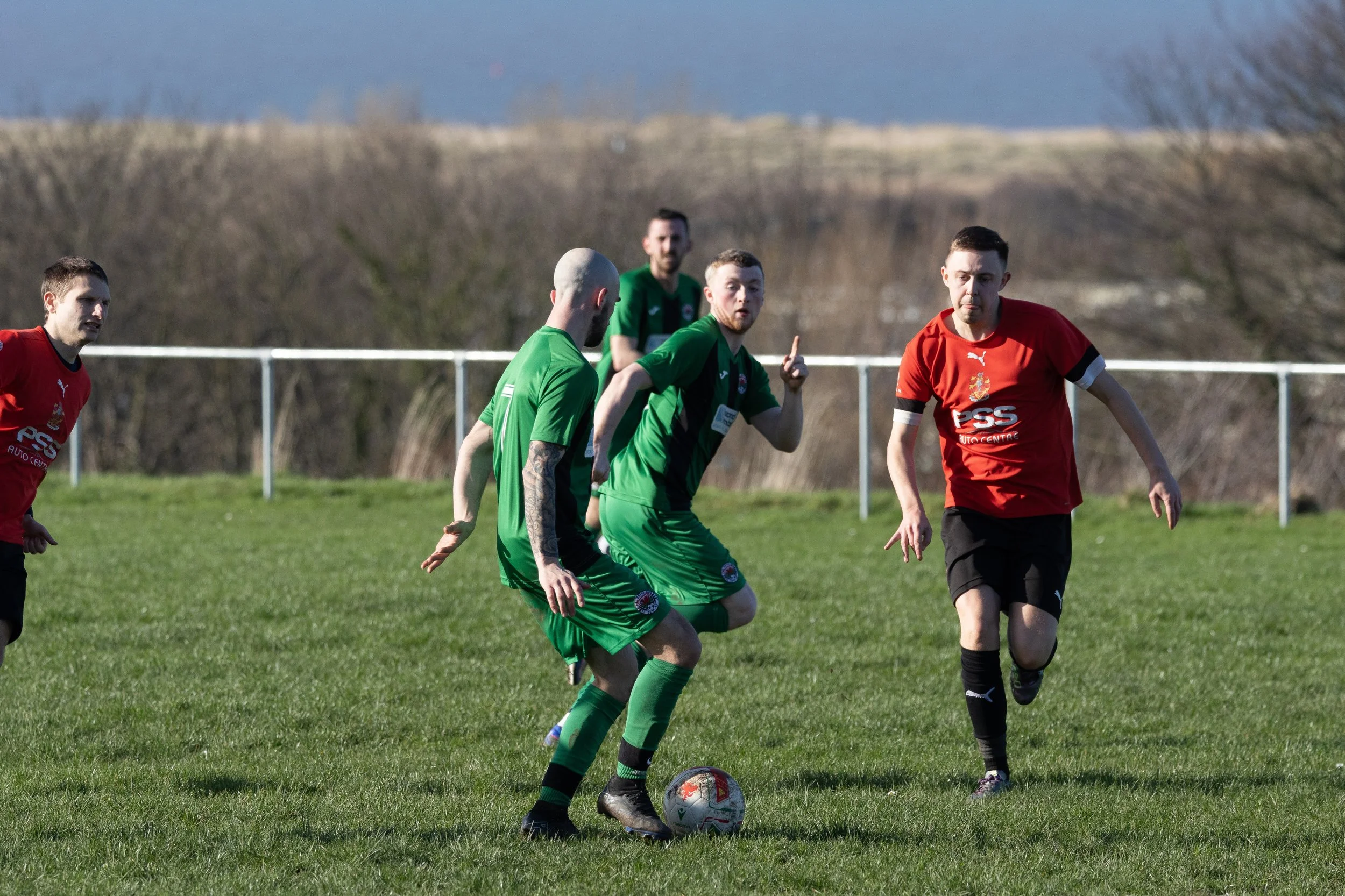 Soccer players on a grassy field during a game, with some wearing red uniforms and others in green, with a background of trees and a distant rail fence.