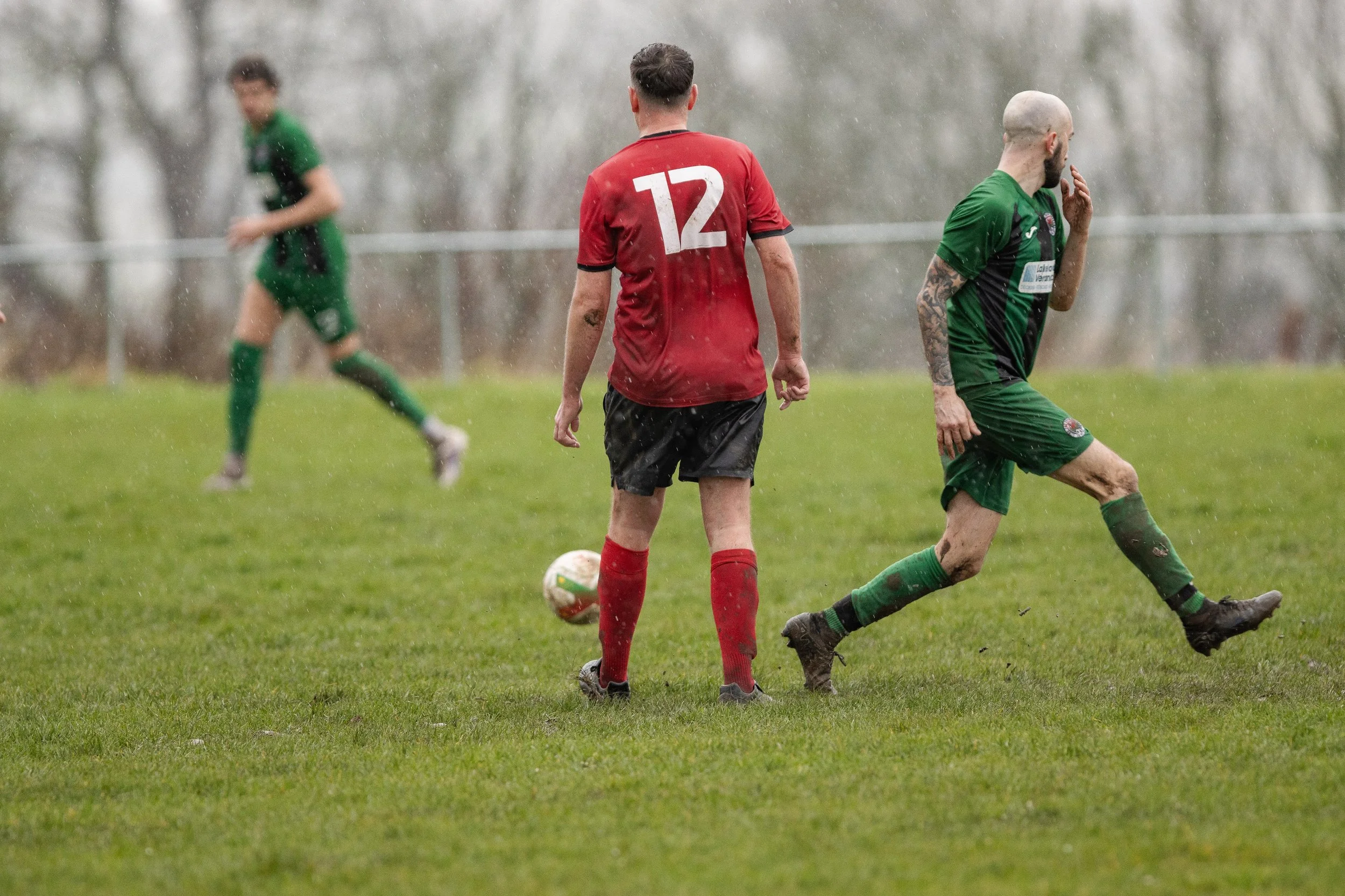 Soccer players in green and red uniforms playing in the rain on a grassy field, with one player in green about to kick the ball and another in red standing nearby.