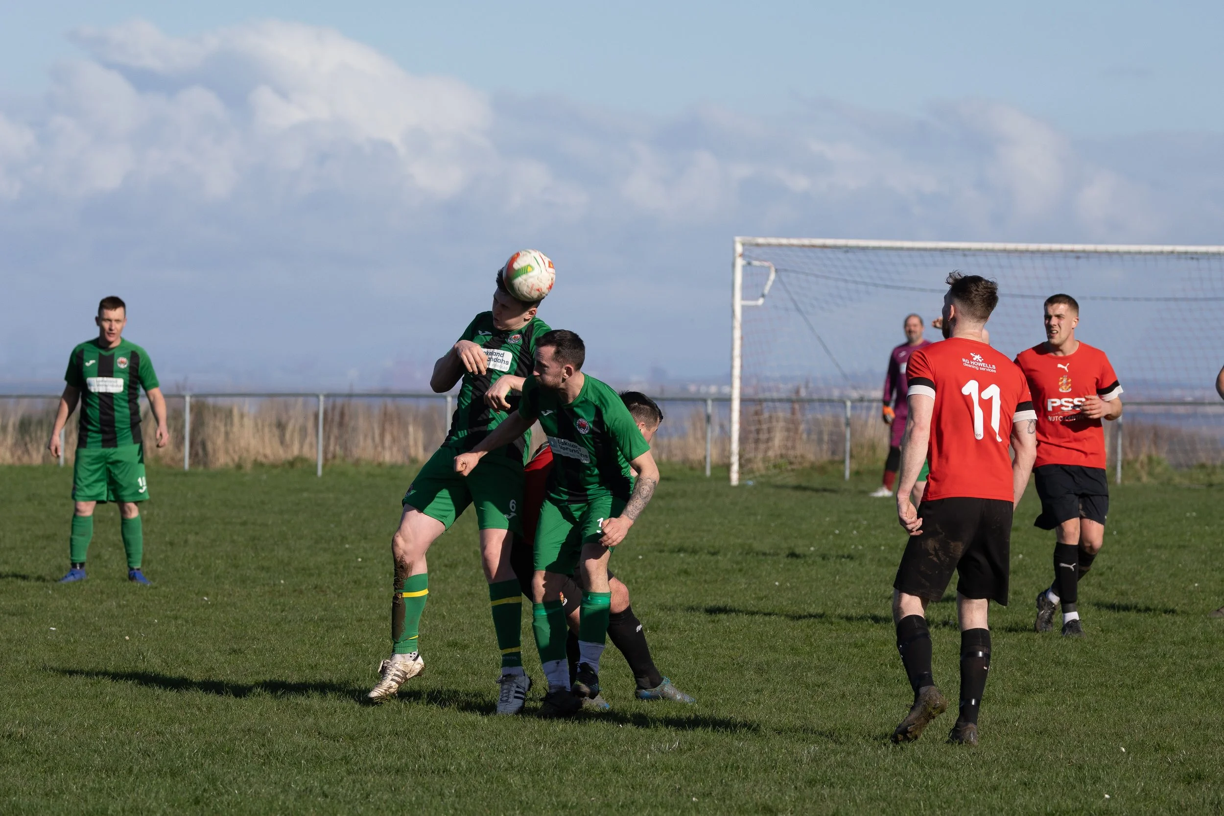 Soccer players competing for the ball on a grassy field, with some players in green uniforms and others in red uniforms, and a goalpost in the background.