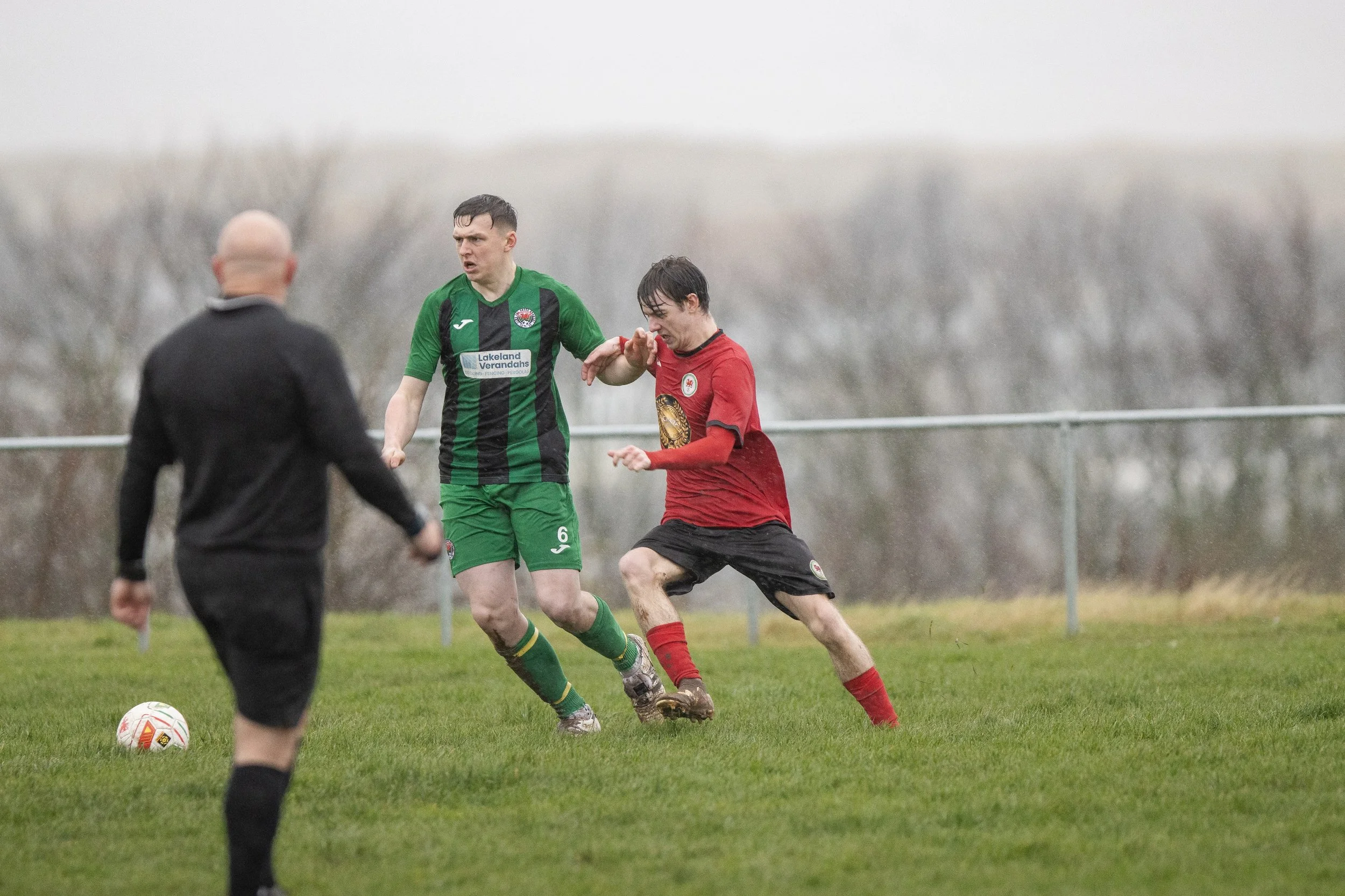 Two soccer players competing for the ball on a grassy field, with a referee in black observing, overcast weather, and trees in the background.