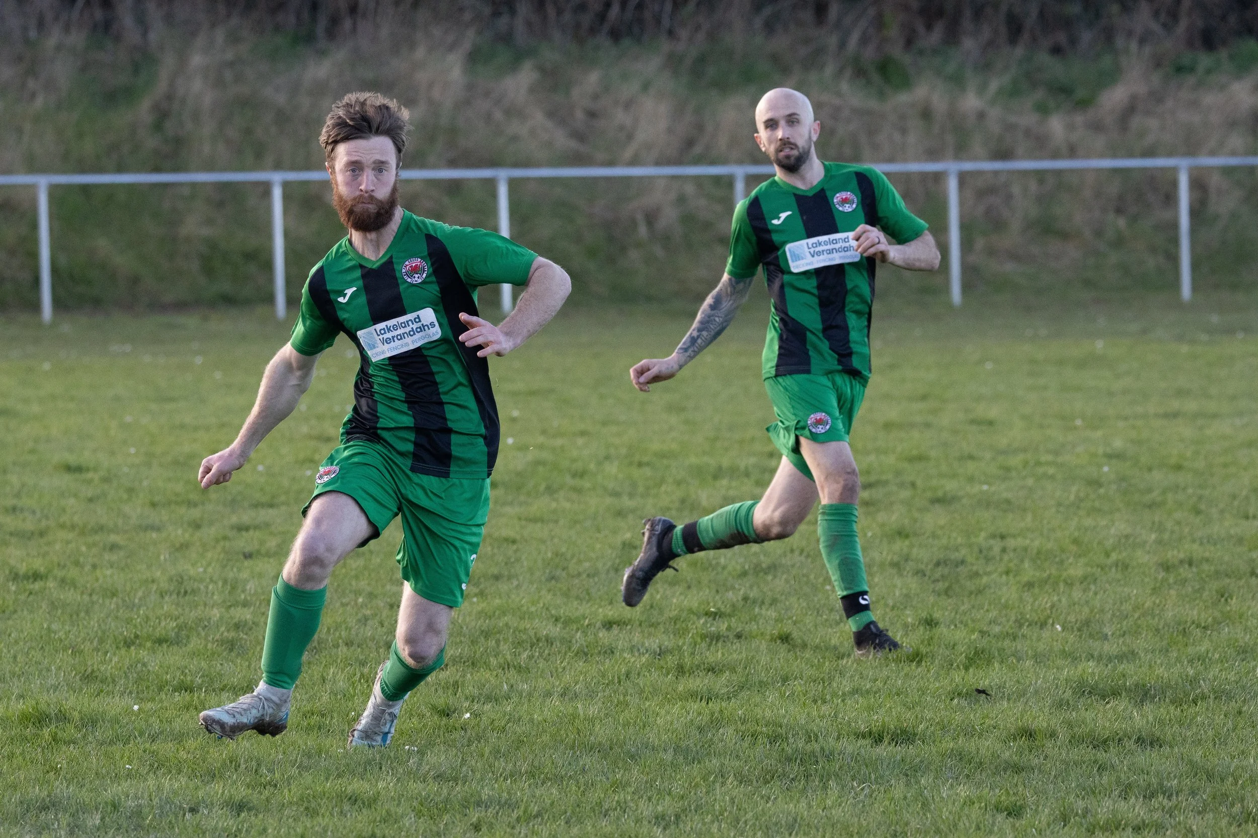 Two male soccer players in green and black uniforms running on a grassy field during a game.