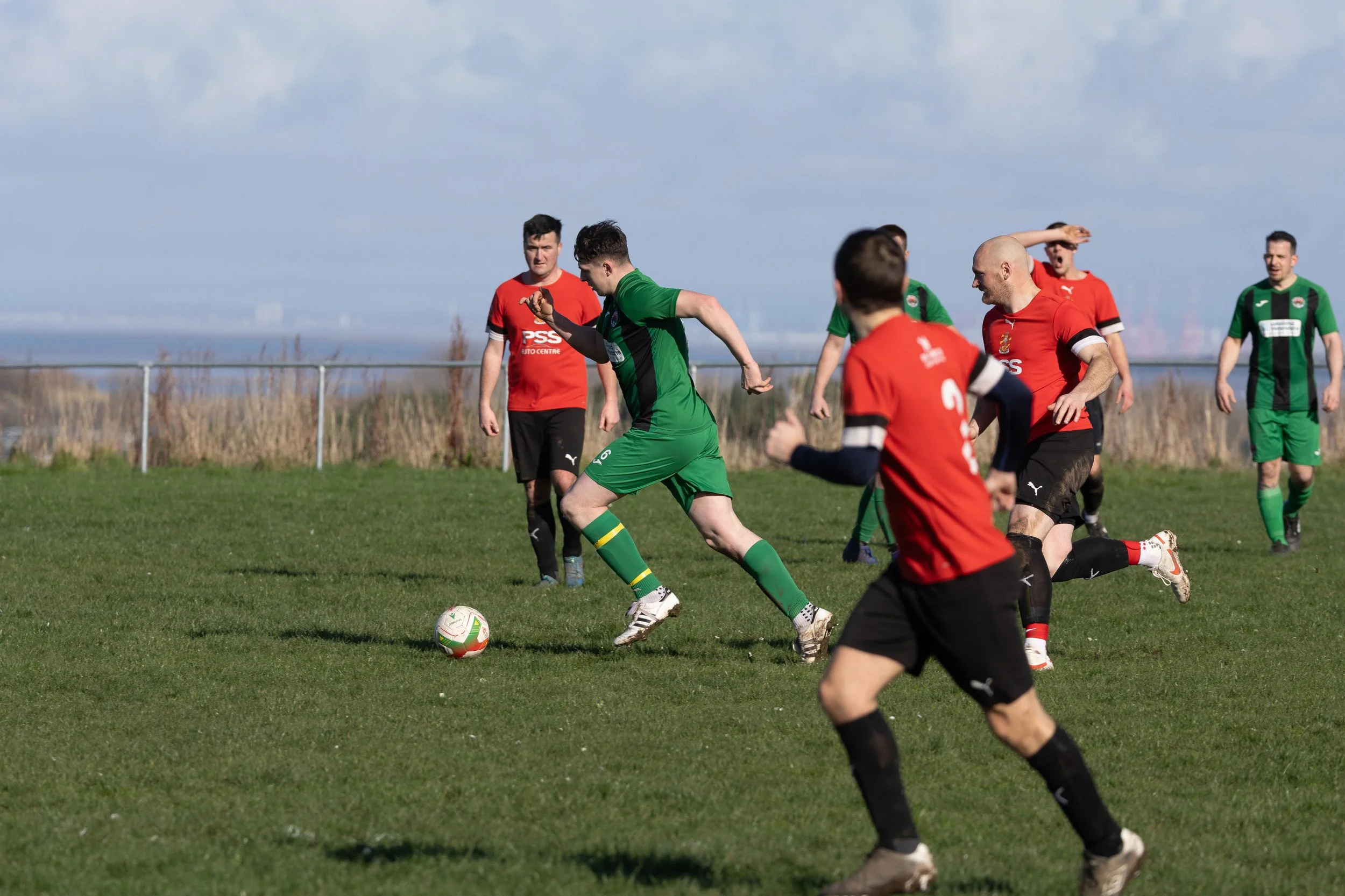 Soccer players competing for the ball during a match on a grass field, with a fence and sky in the background.