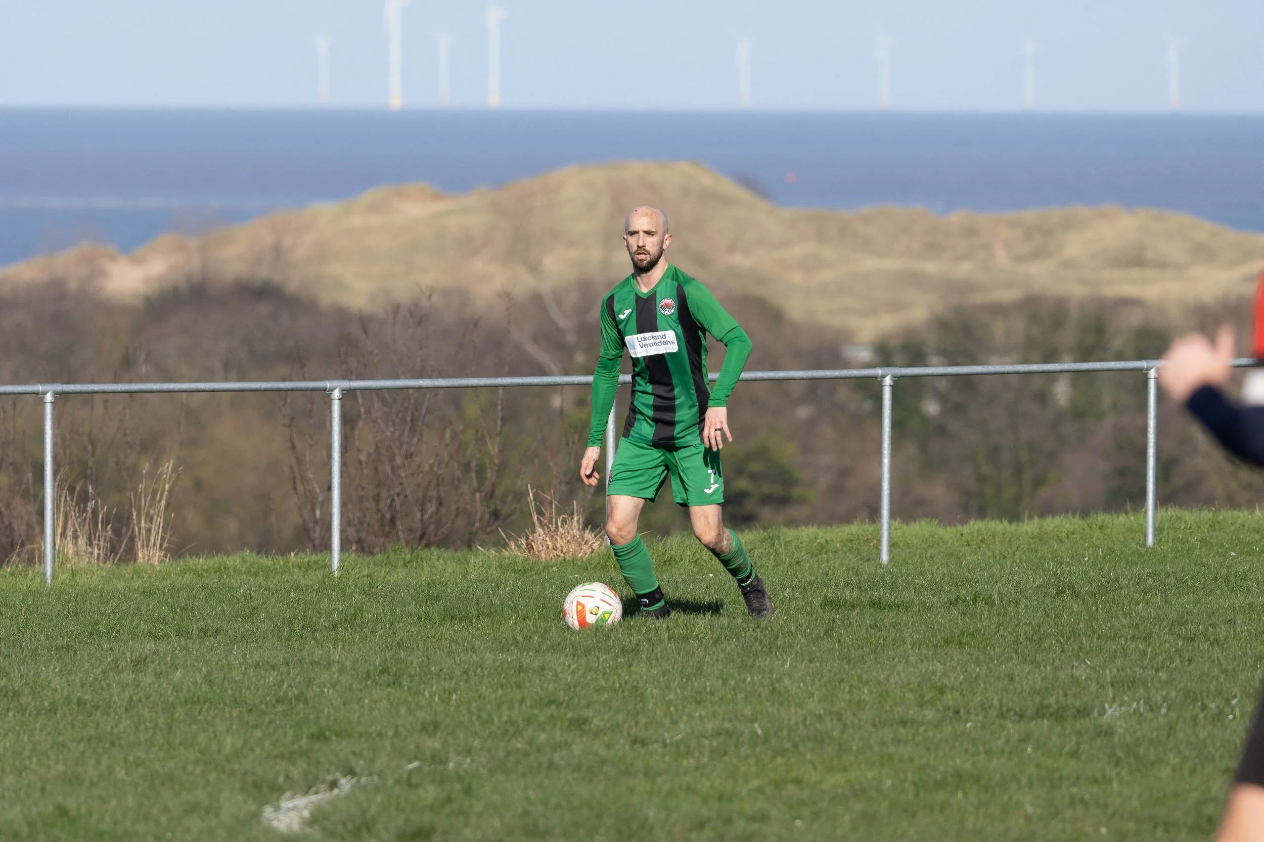 A soccer player in a green and black uniform standing on a grass field near a metal fence, with a landscape of hills and wind turbines in the background.
