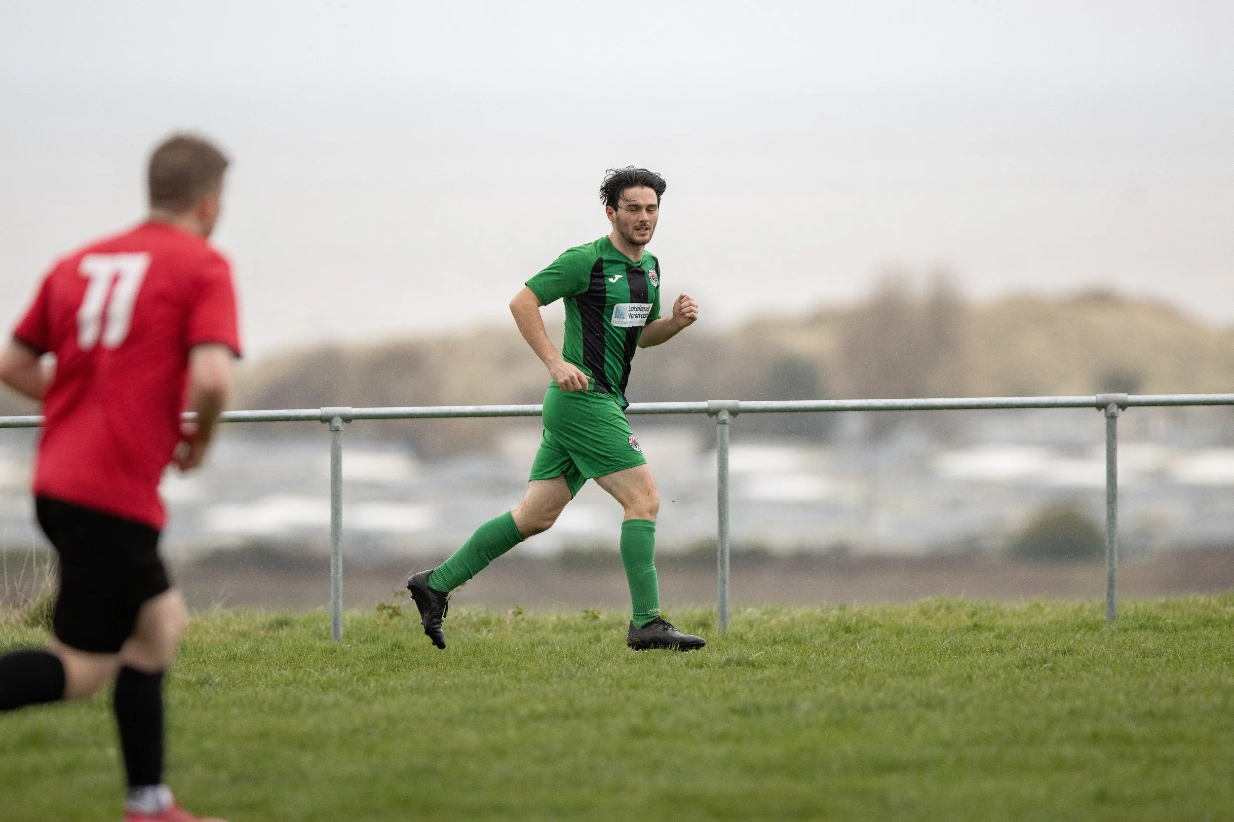 A soccer player in a green and black uniform running on a grassy field during a game, with another player in a red and black uniform partially visible in the foreground.